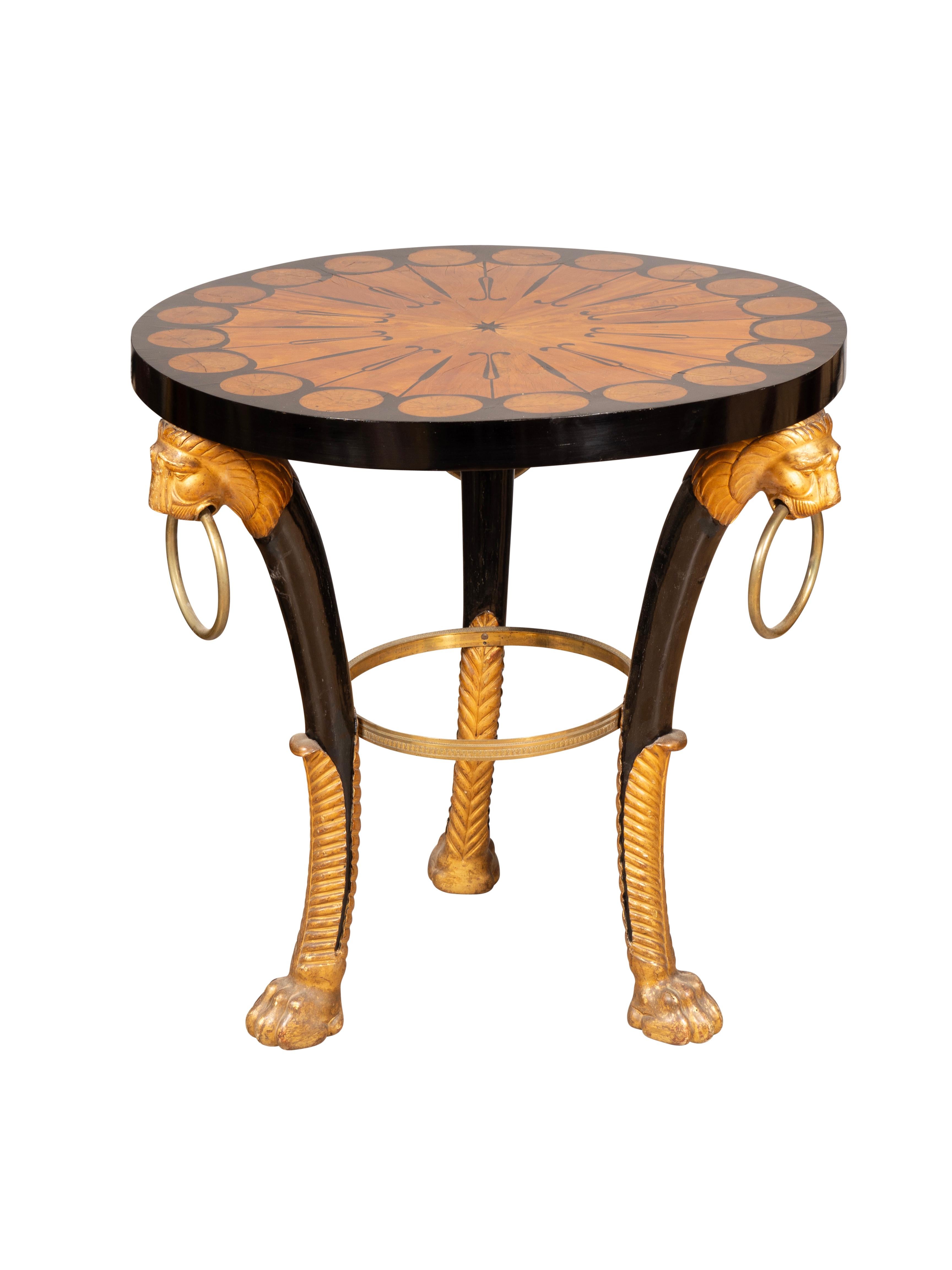 Very much in the manner of Thomas Hope. Circular top with satinwood and ebony inlaid design. The tripartite legs with water gilt lions heads with brass ring handles and ebonized legs joined by a bronze ring above elongated giltwood leaf carved