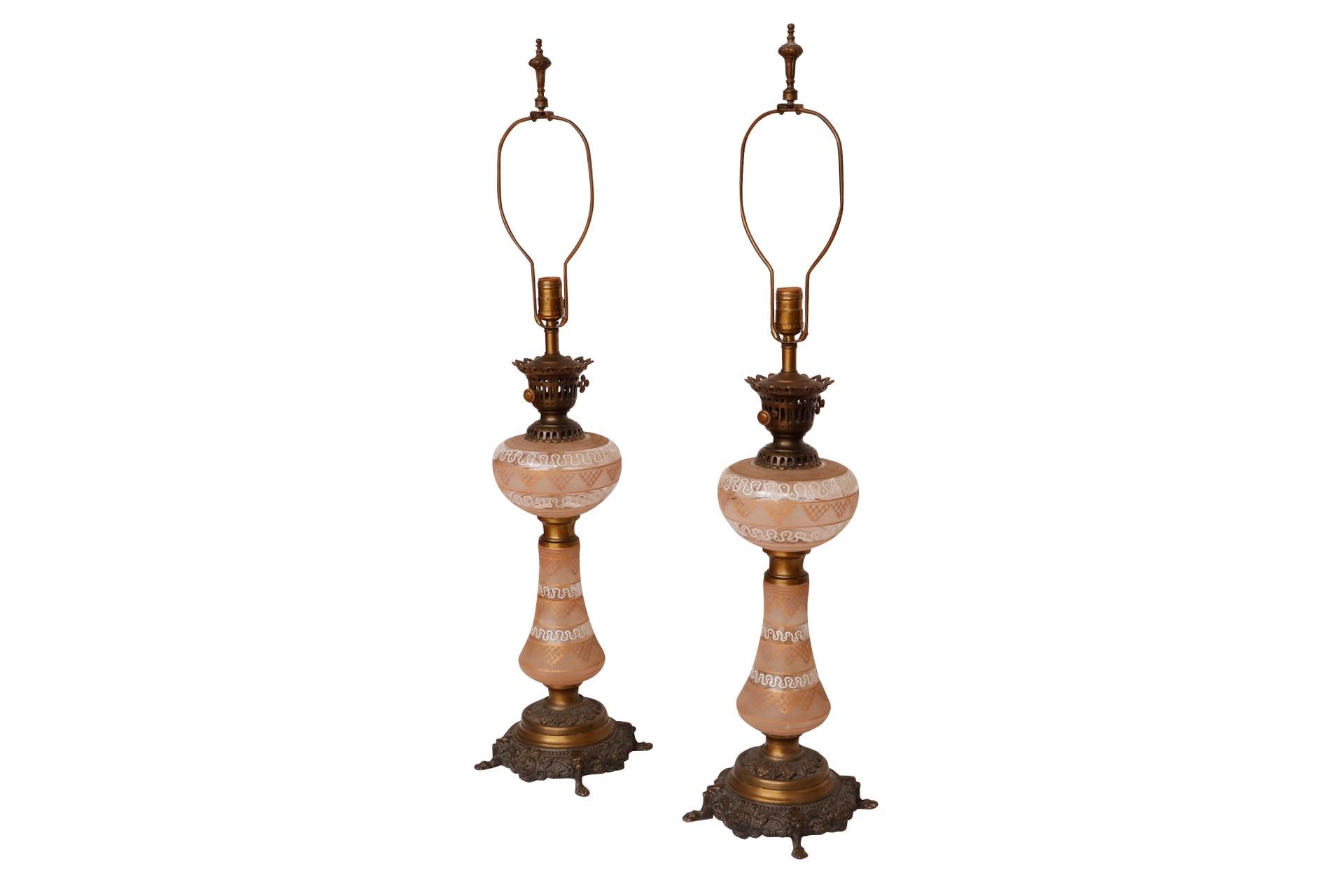 A pair of regency style baluster table lamps made of glass and brass. The glass is decorated with a blush and gold filigree transferware pattern, separated with white serpentine lines. Pierced brass caps and the brass base are pressed with an