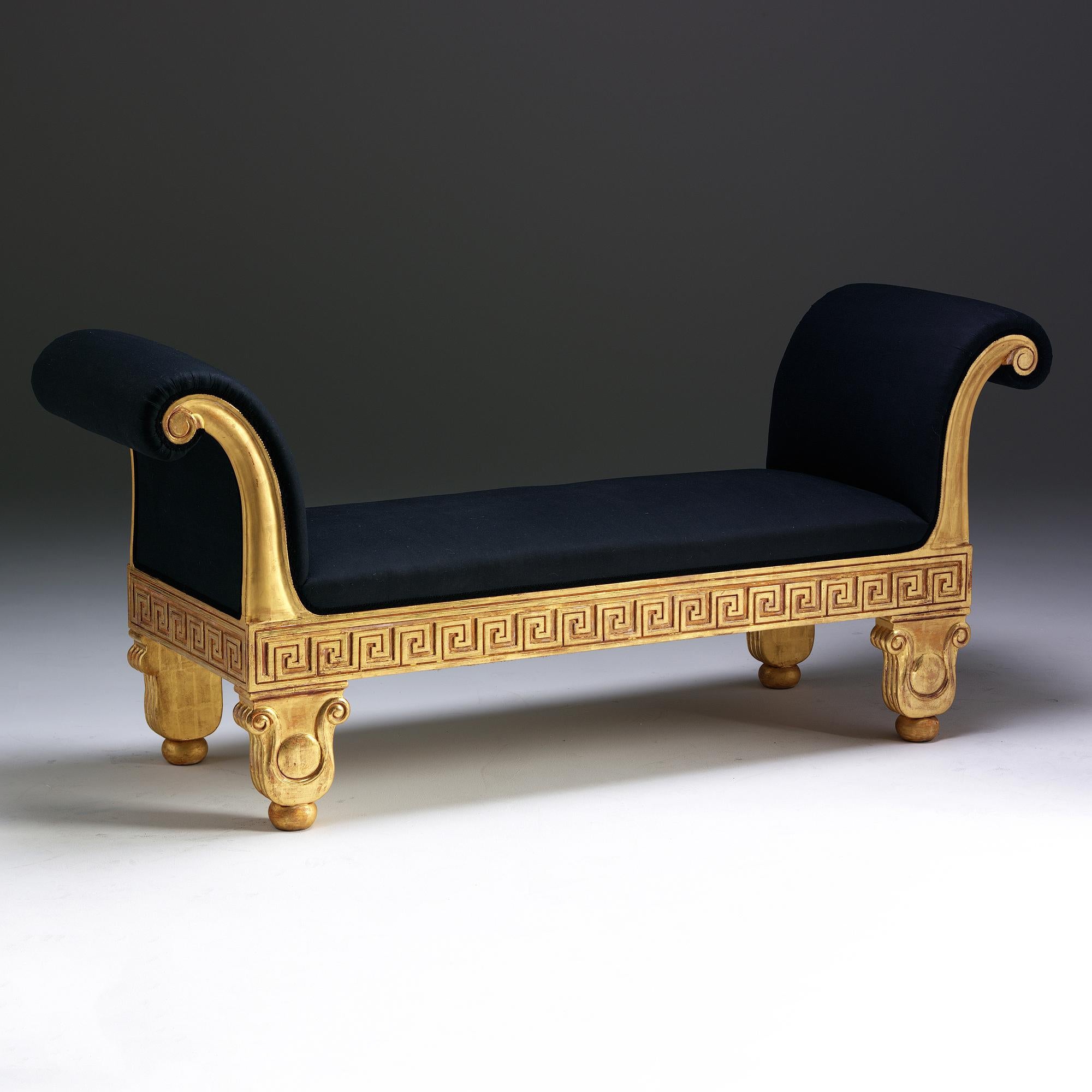 A carved and gilded window seat in the English Regency style. The upholstered and gently curved moulded ends are decorated with carved paterae and rest on Greek key pattern carved rails. The short, lotus leaf shaped legs are centred with paterae and