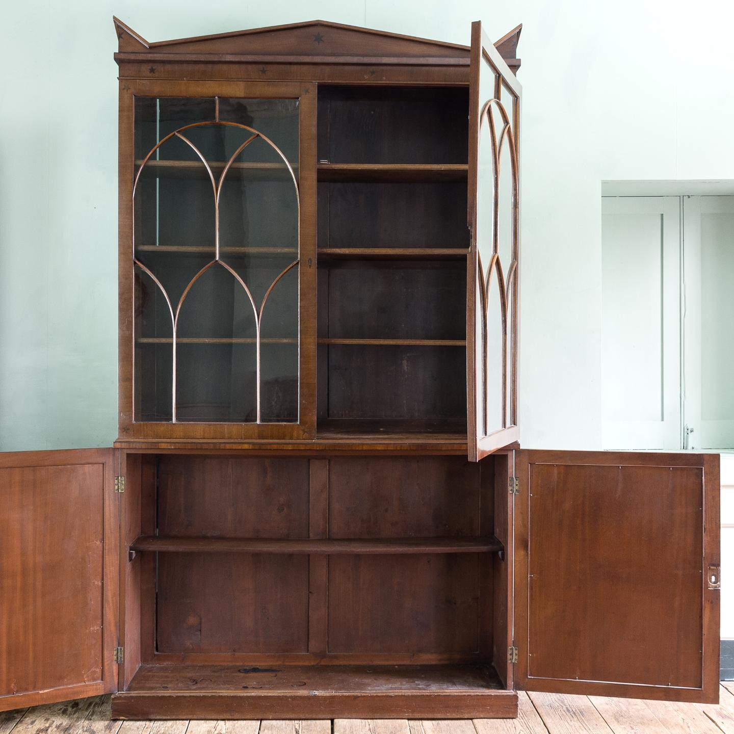 Regency mahogany bookcase, circa 1820, the triangular pediment flanked by acroteria, the Gothic glazed doors enclosing adjustable shelving above base with ebonized inlaid decoration to the doors, on plinth base, with all over inlaid ebonized star