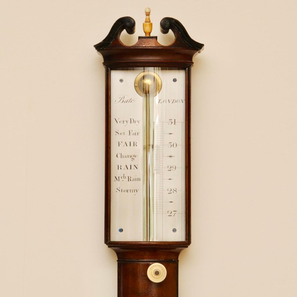 A Regency period mahogany and ebony inlaid bow fronted stick barometer with swan neck pediment above silvered scales that are signed Bate, London; the flame mahogany case with a long elegant thermometer and an urn cistern cover with ebony fluted