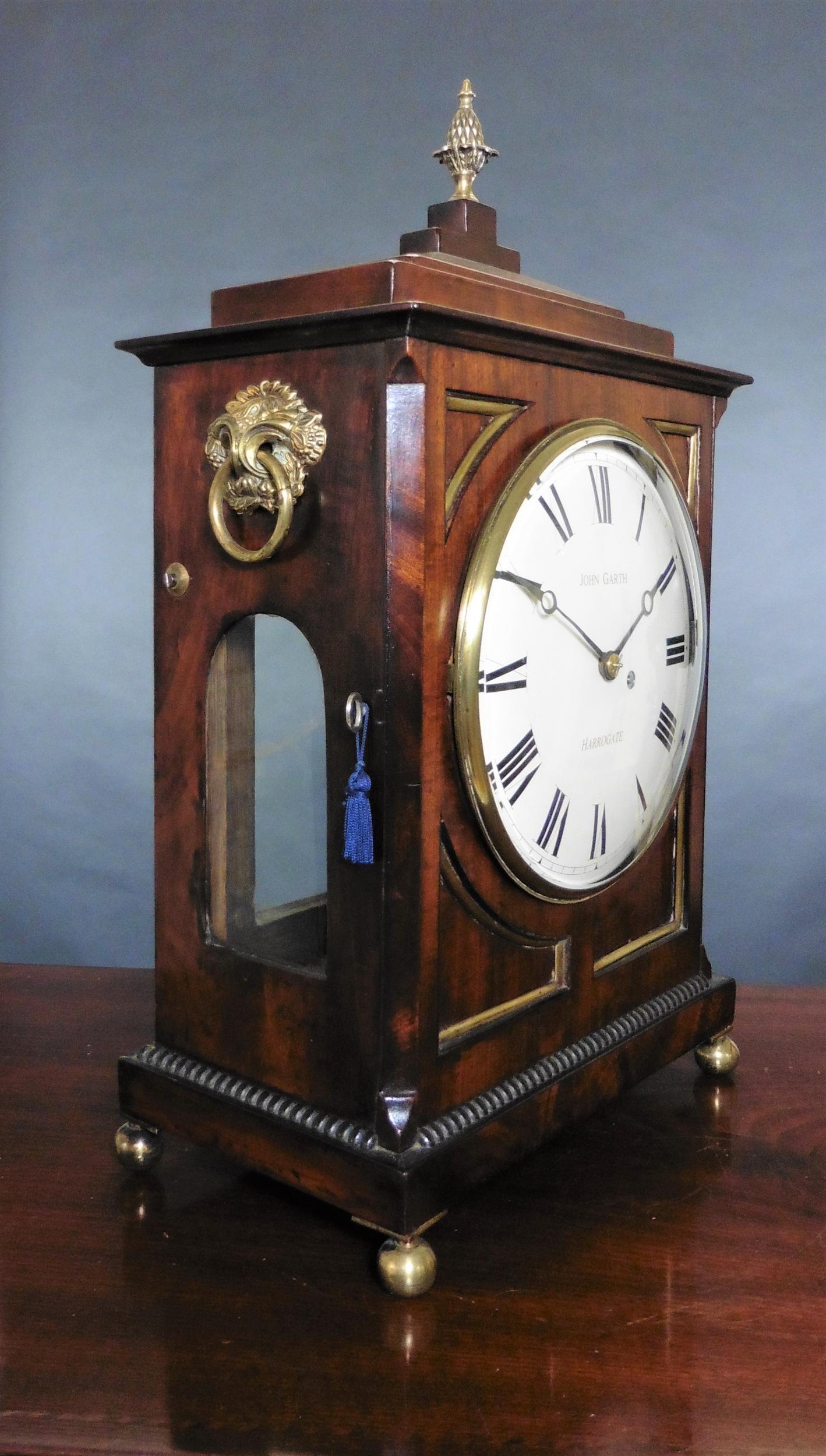 Regency bracket clock by
John Garth, Harrogate

Mahogany case standing on a stepped base with ribbed decoration and standing on four brass ball feet, chamfered top surmounted by a brass pineapple finial.

Cast brass bezel with bevelled glass
