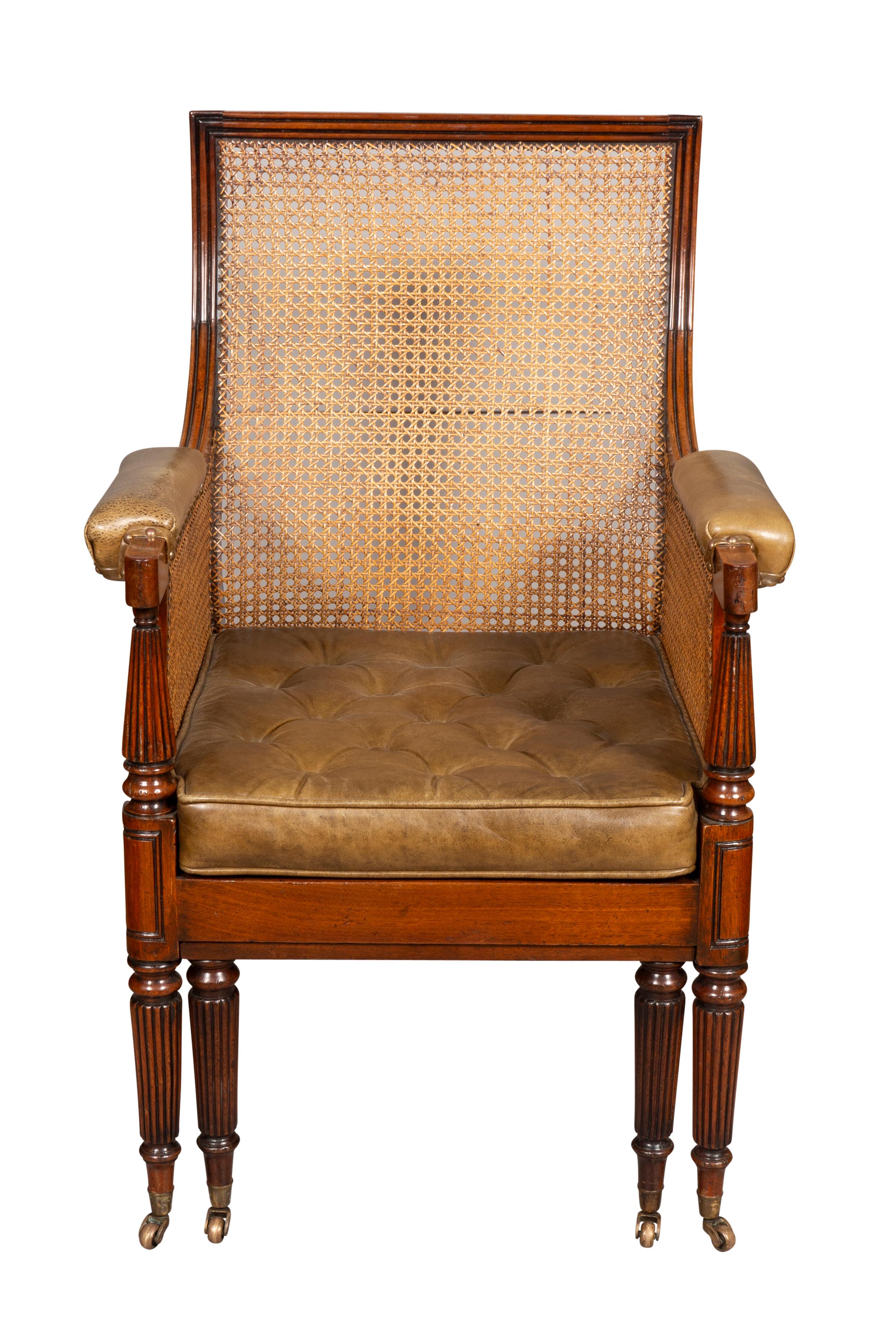 With straight molded crestrail over a caned back and sides, the arms with leather arm pads, tufted green leather seat cushion over a caned seat. Pull out foot rest. Raised on circular tapered reeded legs with brass cup casters.