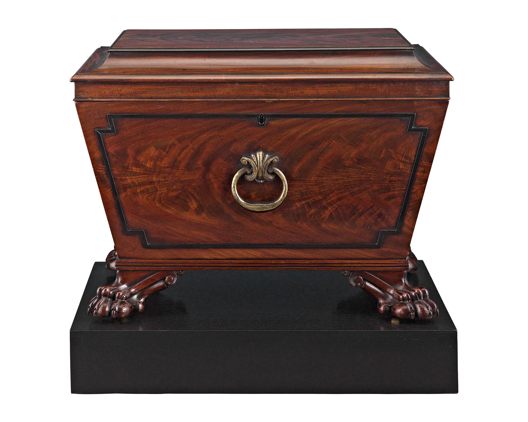 This refined Regency-period wine cellarette is crafted of rich mahogany and boasts beautifully carved details. The sarcophagus form is distinguished by ebonized accents and handsome lion paw feet. Complete with its original brass side handles and