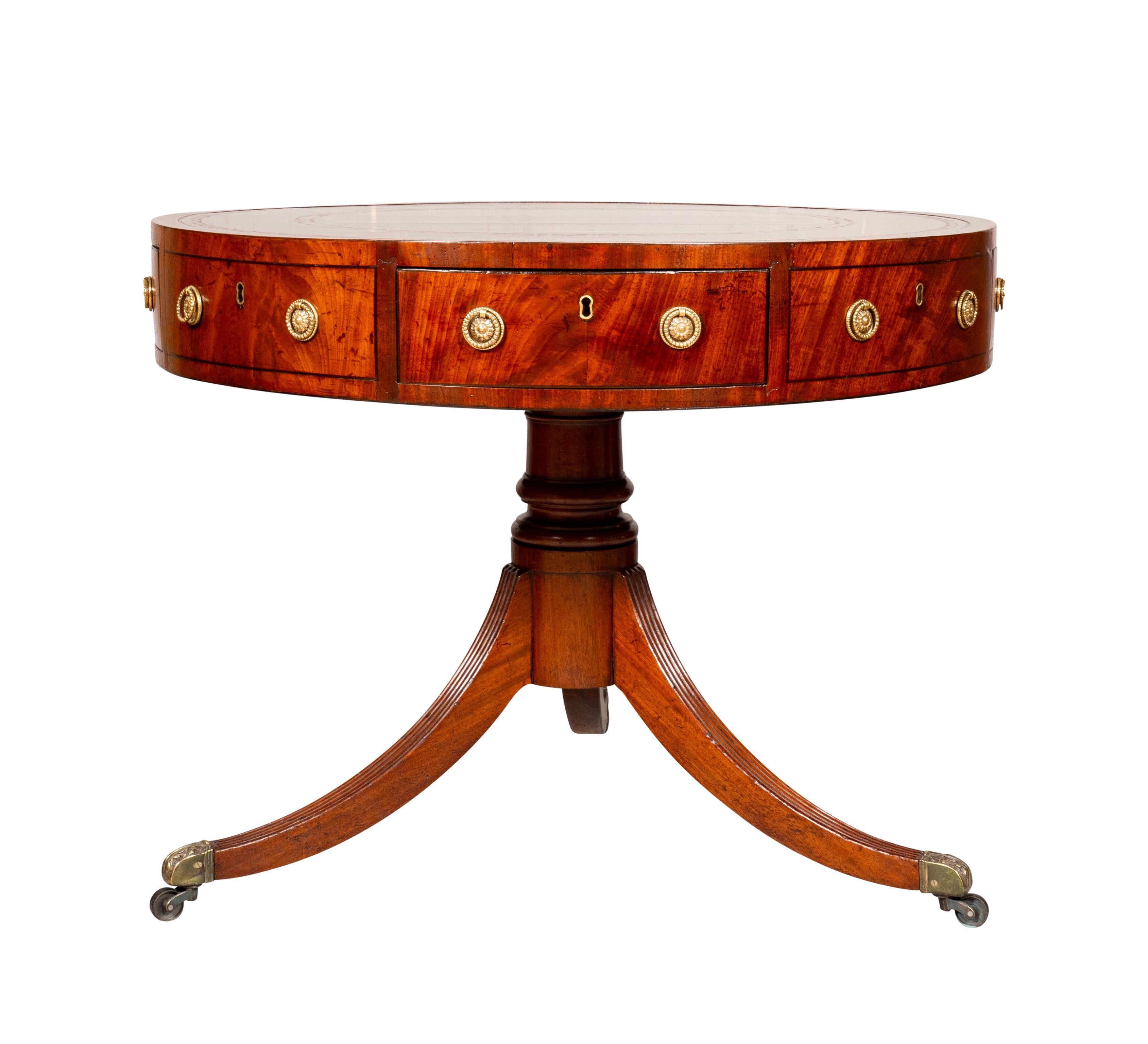Circular cross banded top with brown tooled leather .The frieze containing drawers. Turned support joining three saber legs with casters.