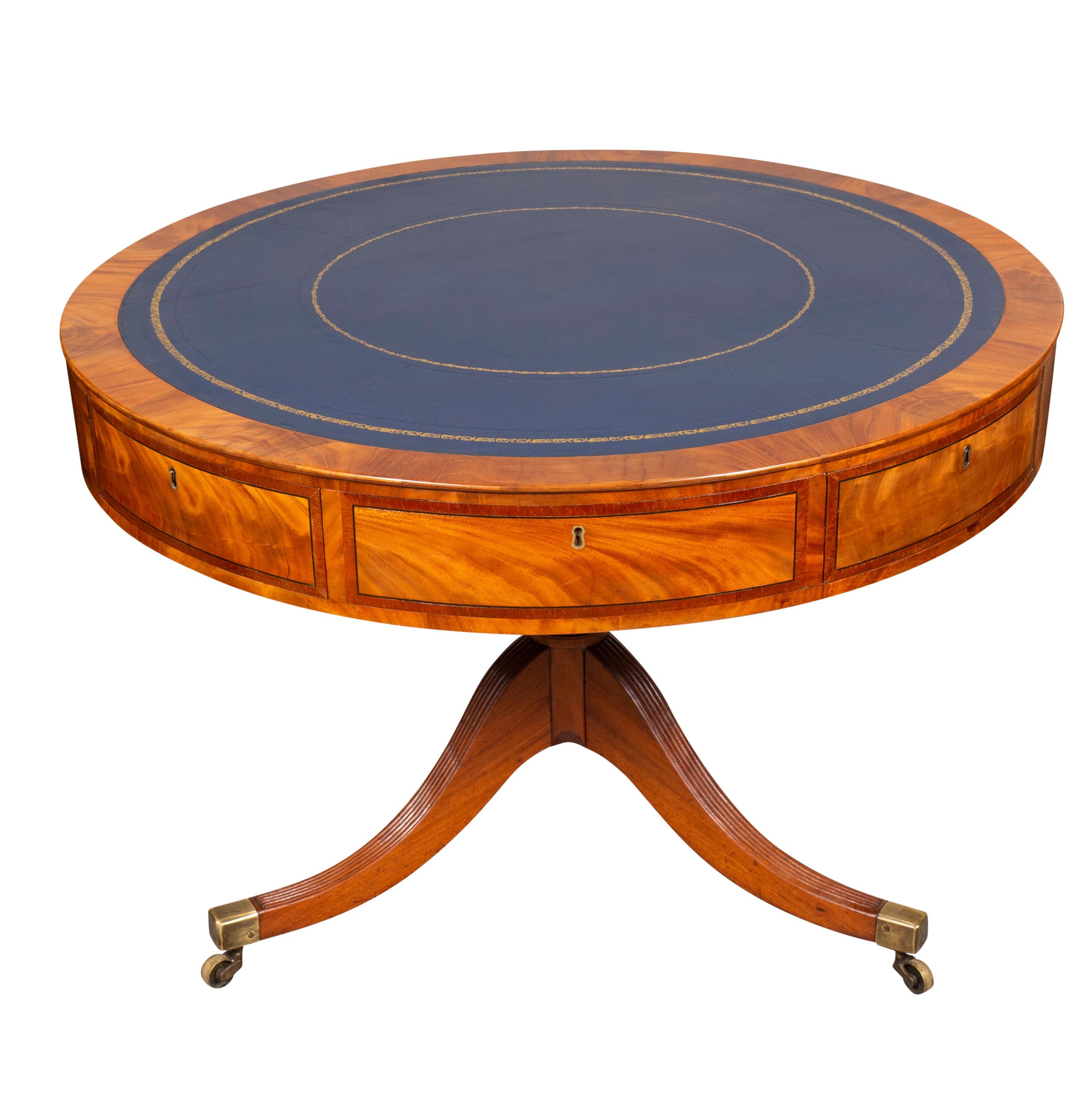 Circular with blue leather top with cross banded edge. The frieze containing drawers, all on a turned supported joining three reeded saber legs with casters.