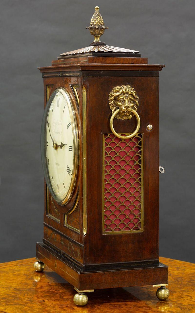 Regency mahogany bracket clock with chamfer top, finely figured brass bound case with side carrying handles, side fishscale sound frets surmounted by a pineapple finial and standing on brass ball feet.
 
Painted dial with Roman numerals, original