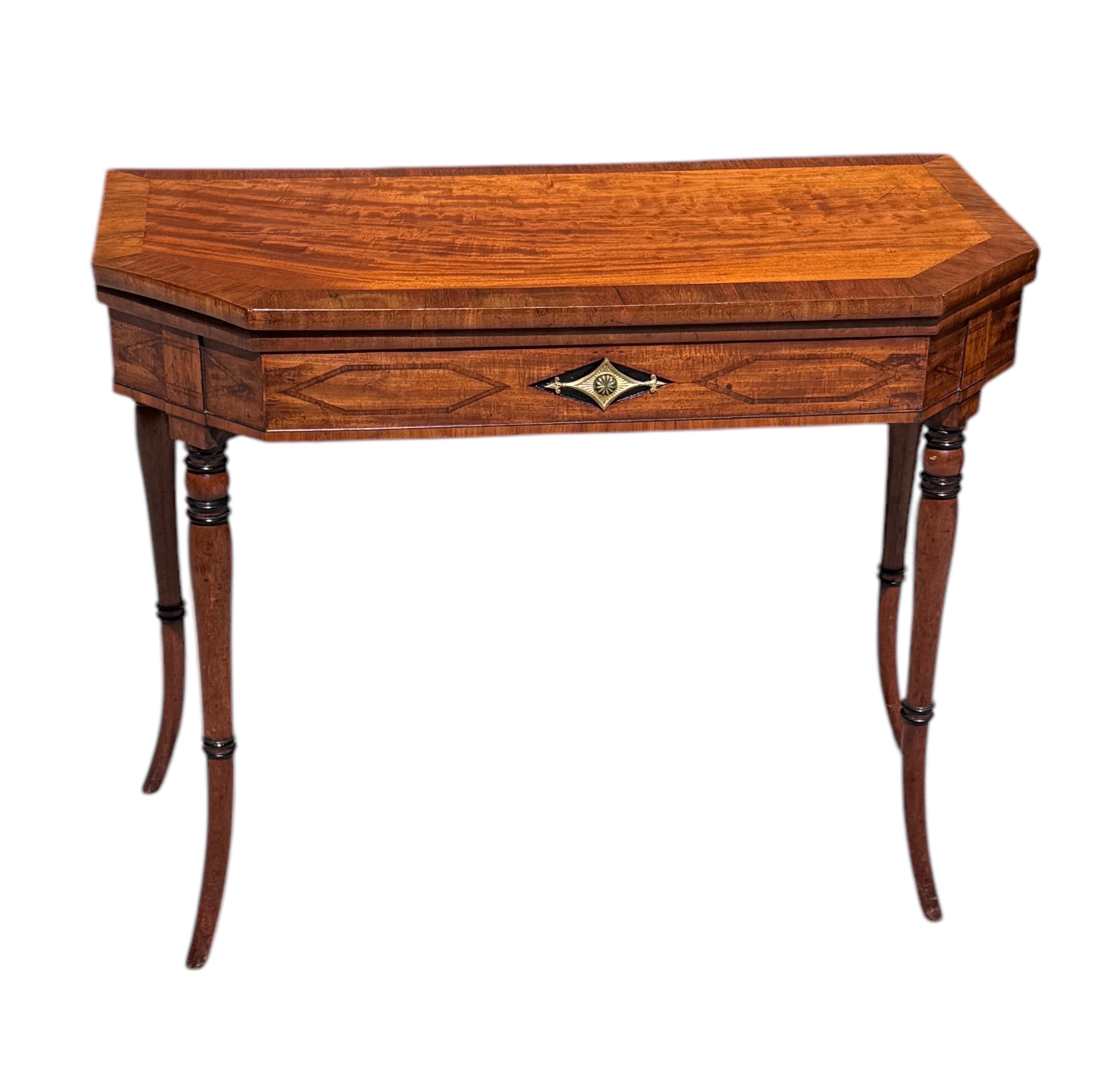 Mesa de juegos de caoba Regency,  con parte superior de cuero
Patas de caoba torneadas, ahusadas y con collar, asentadas sobre 
Friso de caoba con incrustaciones geométricas, con una montura de latón sobre fondo ebonizado debajo de una tapa de caoba