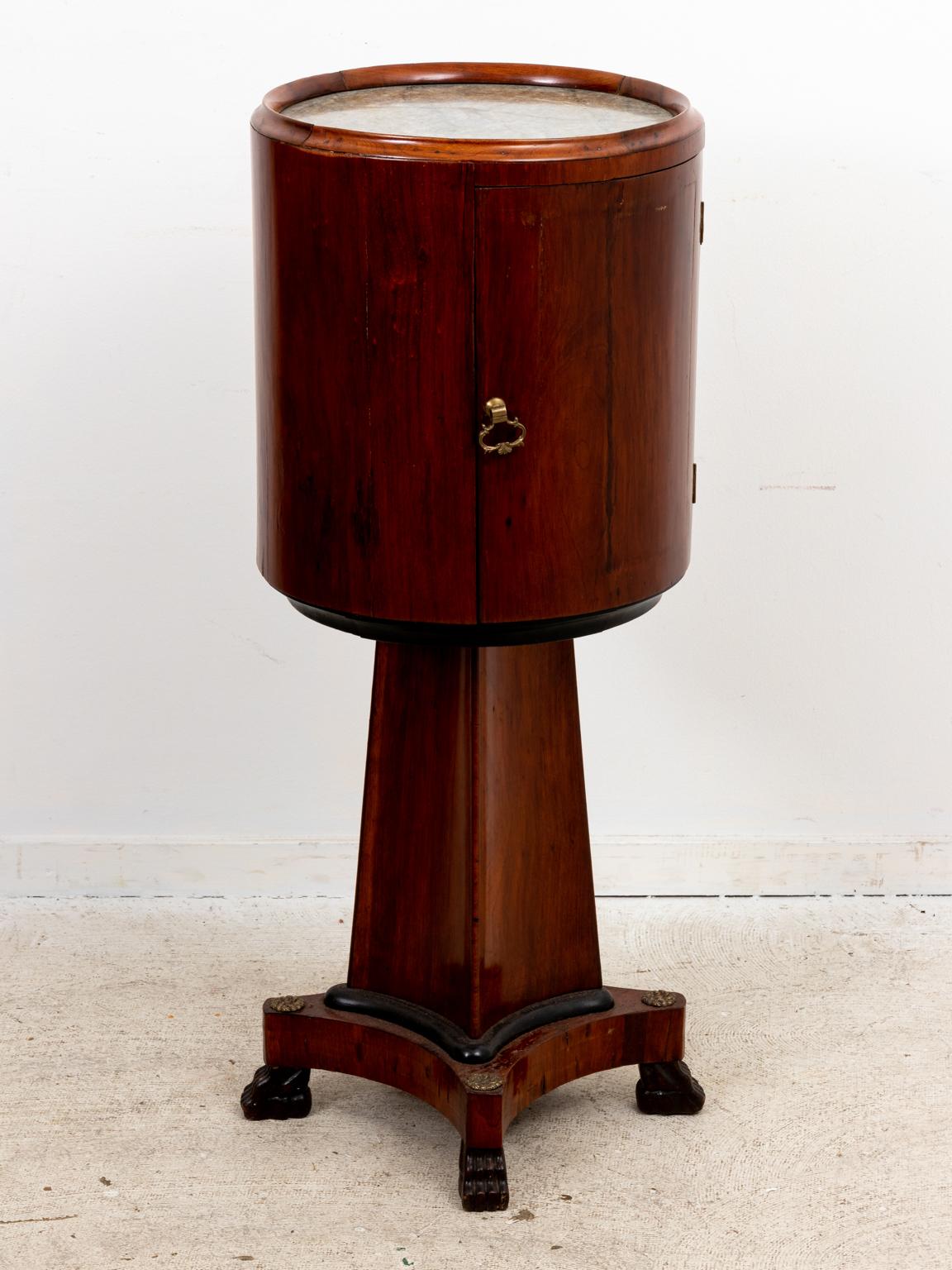 Regency style round mahogany bedside table with marble top and metal hardware on the curved front cabinet and bottom drawer. The base features a tapered pedestal on four carved feet. Please note of wear consistent with age including patina to the