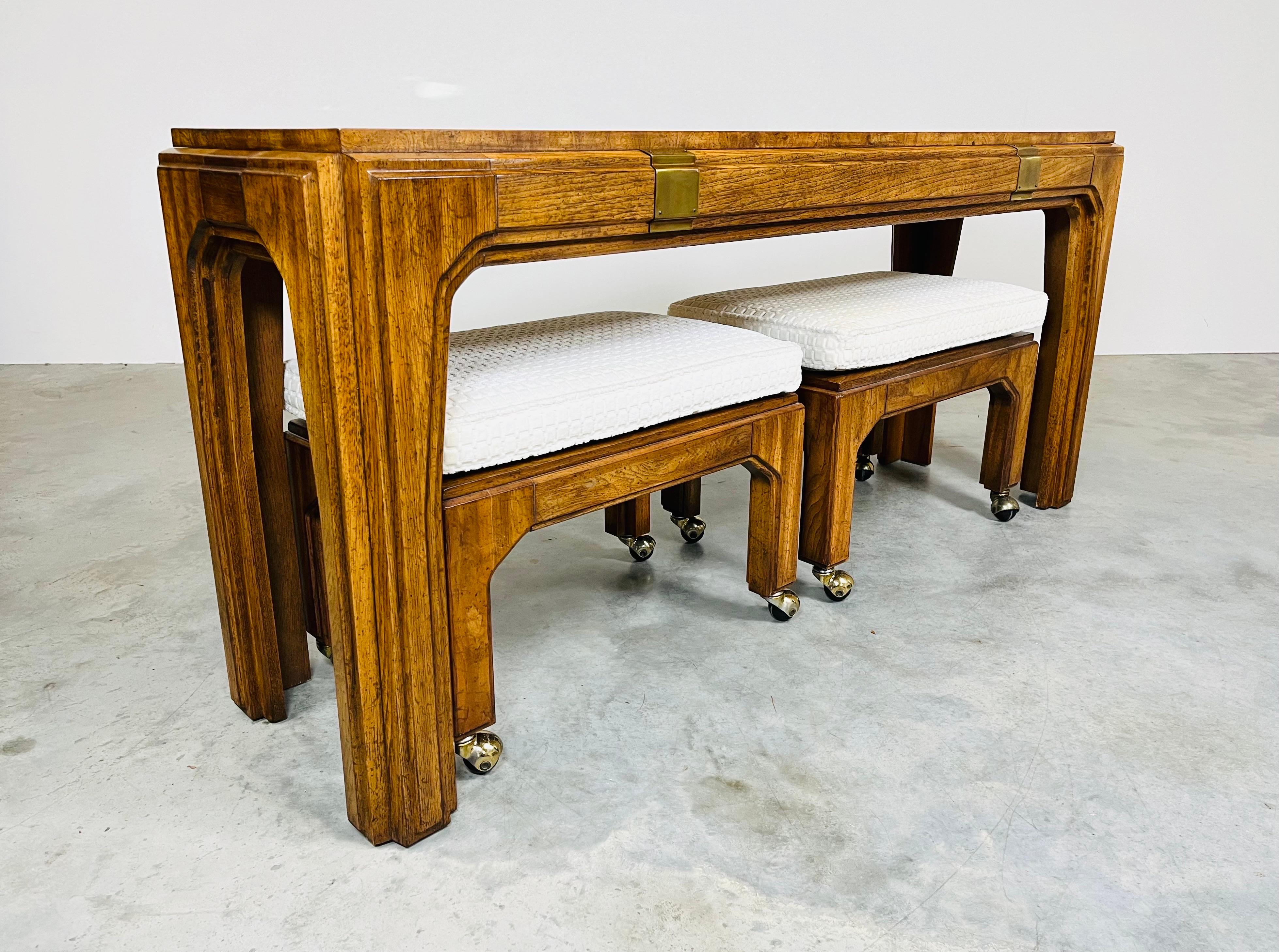 A beautiful 3-piece Hollywood Regency console table having matching stow away benches with detachable cushions on ball casters attributed to Henredon. The console table and benches are constructed of solid mahogany with olive wood center top on the