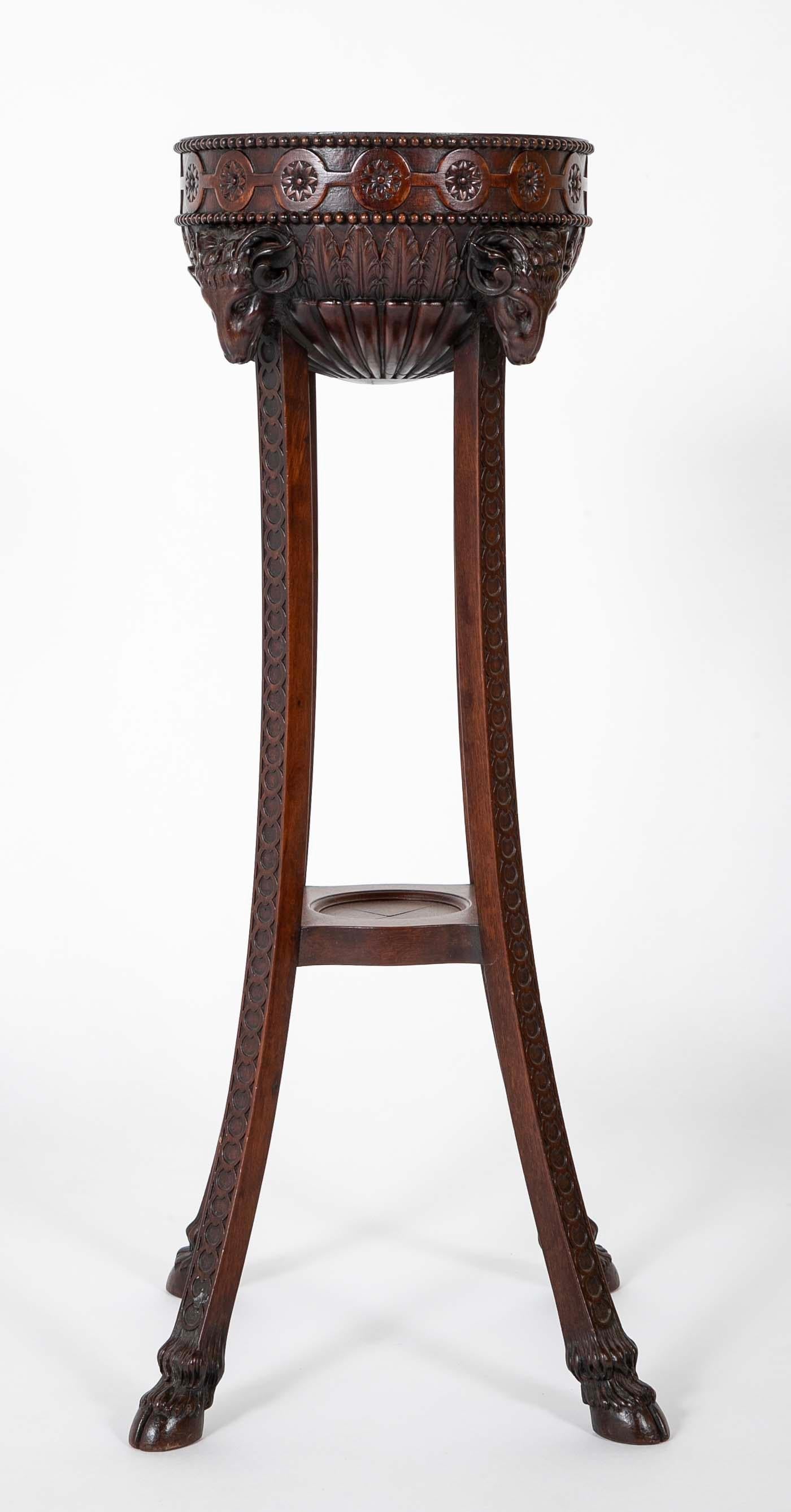 English Regency style carved mahogany plant stand with four splayed legs supporting a central bowl decorated with four ram heads. The bowl with beaded rim and bands of acanthus leaves in relief. The legs decorated with interlocking circles ending in