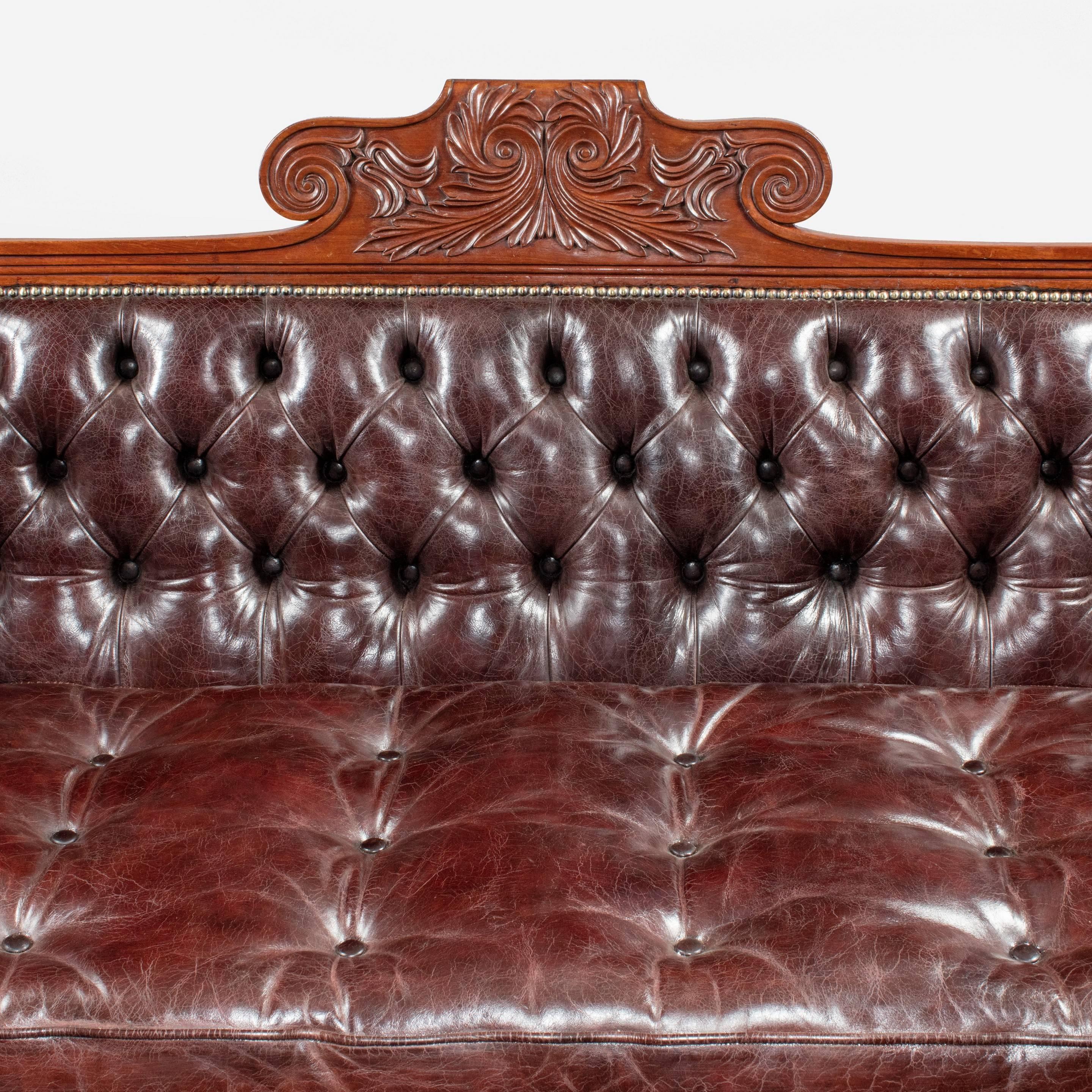 A late Regency mahogany sofa, boldly carved with roundels, the curved cresting centered on a scrolling motif and supported on lyre fronted legs with the original castors, re-upholstered in deep-buttoned burgundy leather.