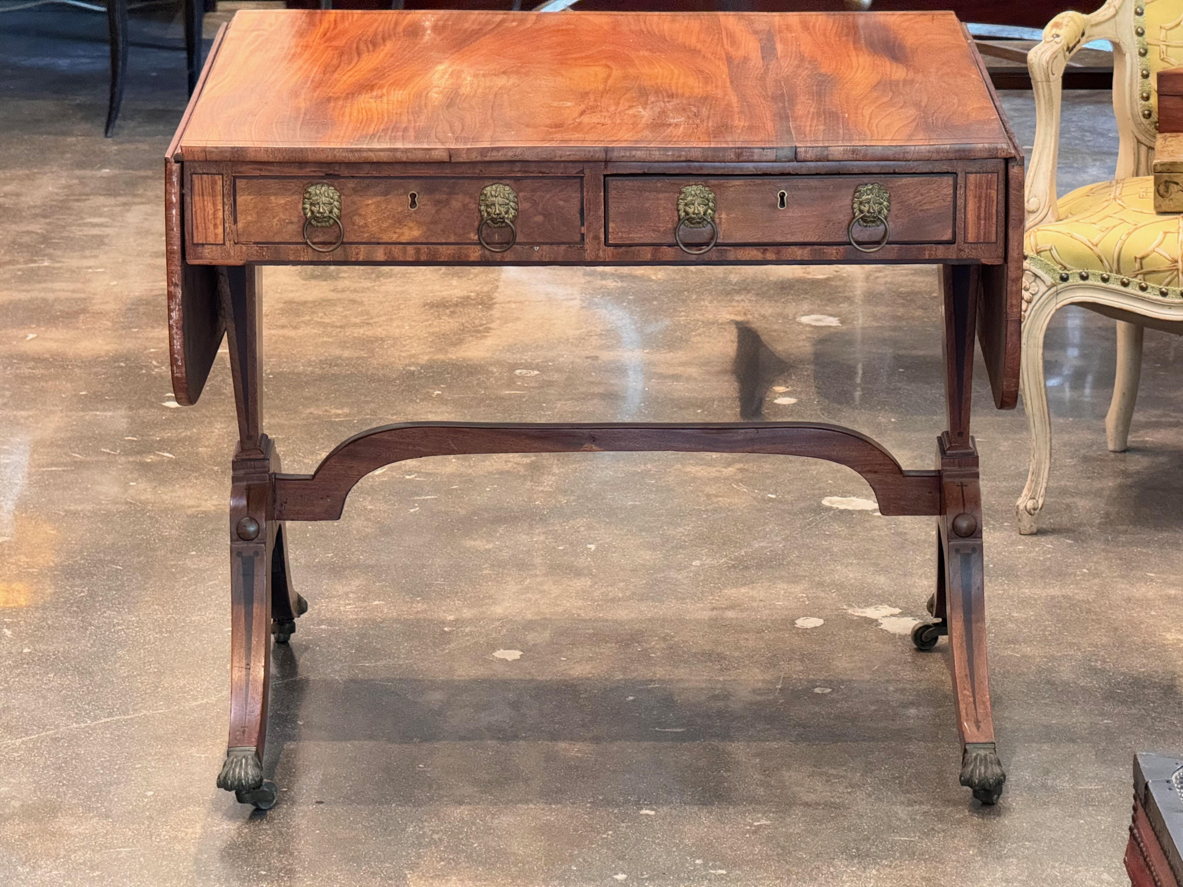 Early 19th century English Regency sofa table in mahogany, featuring two drop leaves and two drawers adorned with brass lion mask pulls. The top displays inlaid corner flourishes and fine grain patterning. Supported by splayed legs joined by a