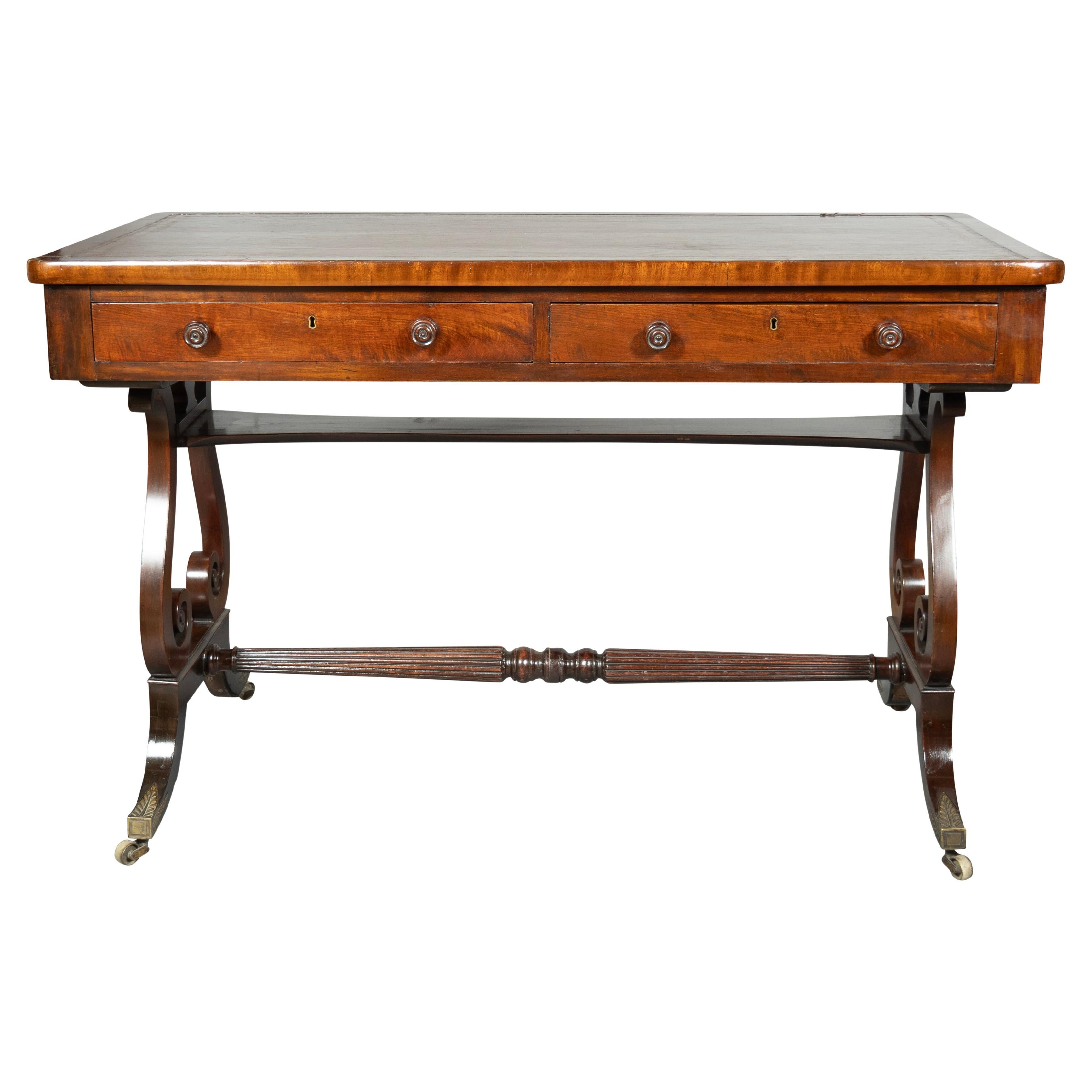With an old brown leather inset top with banded edge over two drawers and raised on a trestle base with Lyre ends and turned stretcher, saber legs and pointed leaf tip caster cups and wheels.