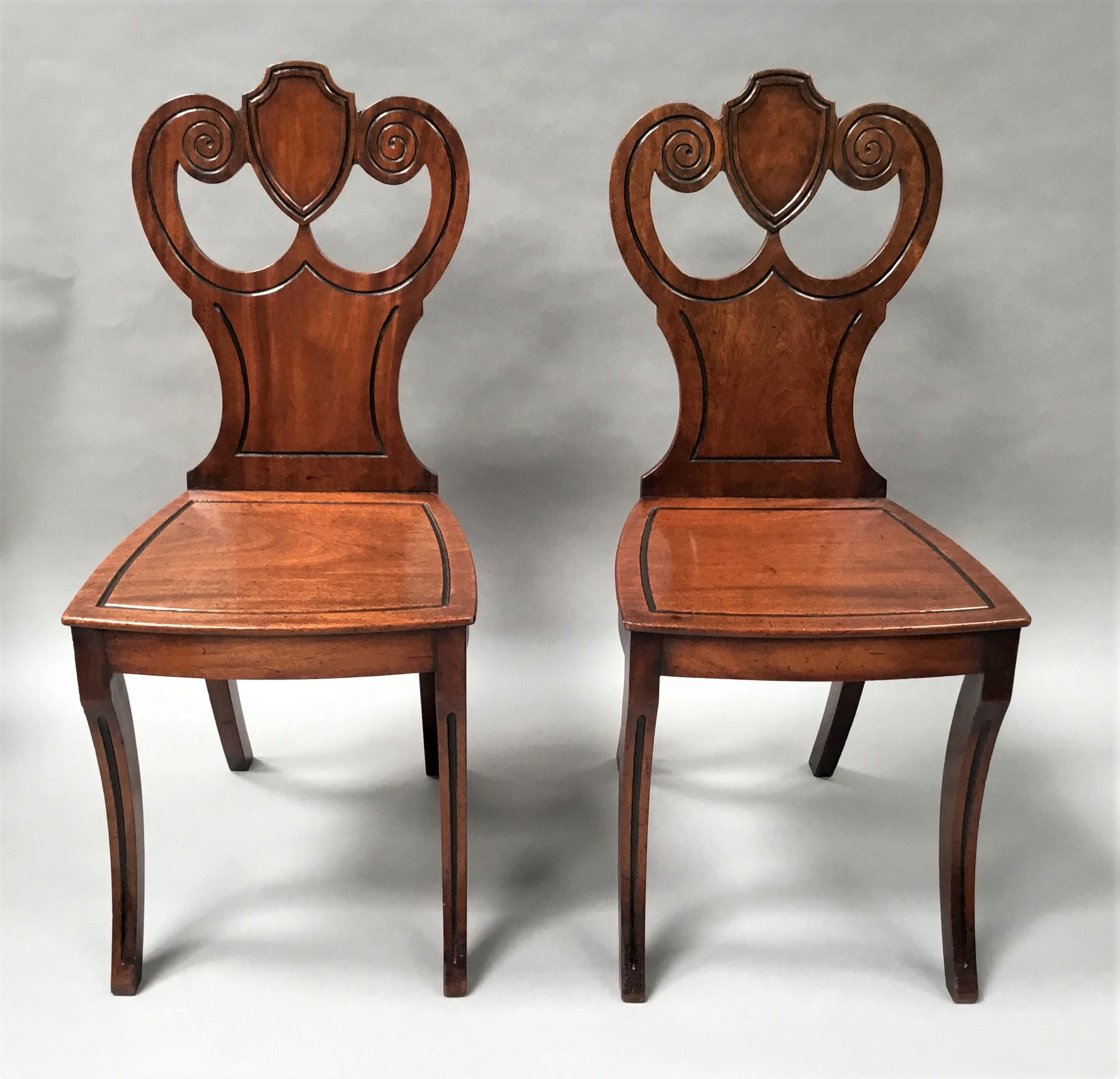 Good Regency pair of mahogany hall chairs of stylish design, the waisted shaped backs headed with a central shield flanked by open scrolls all with ebonised single fluted decoration in a scroll work design. The bow fronted shaped seats with the