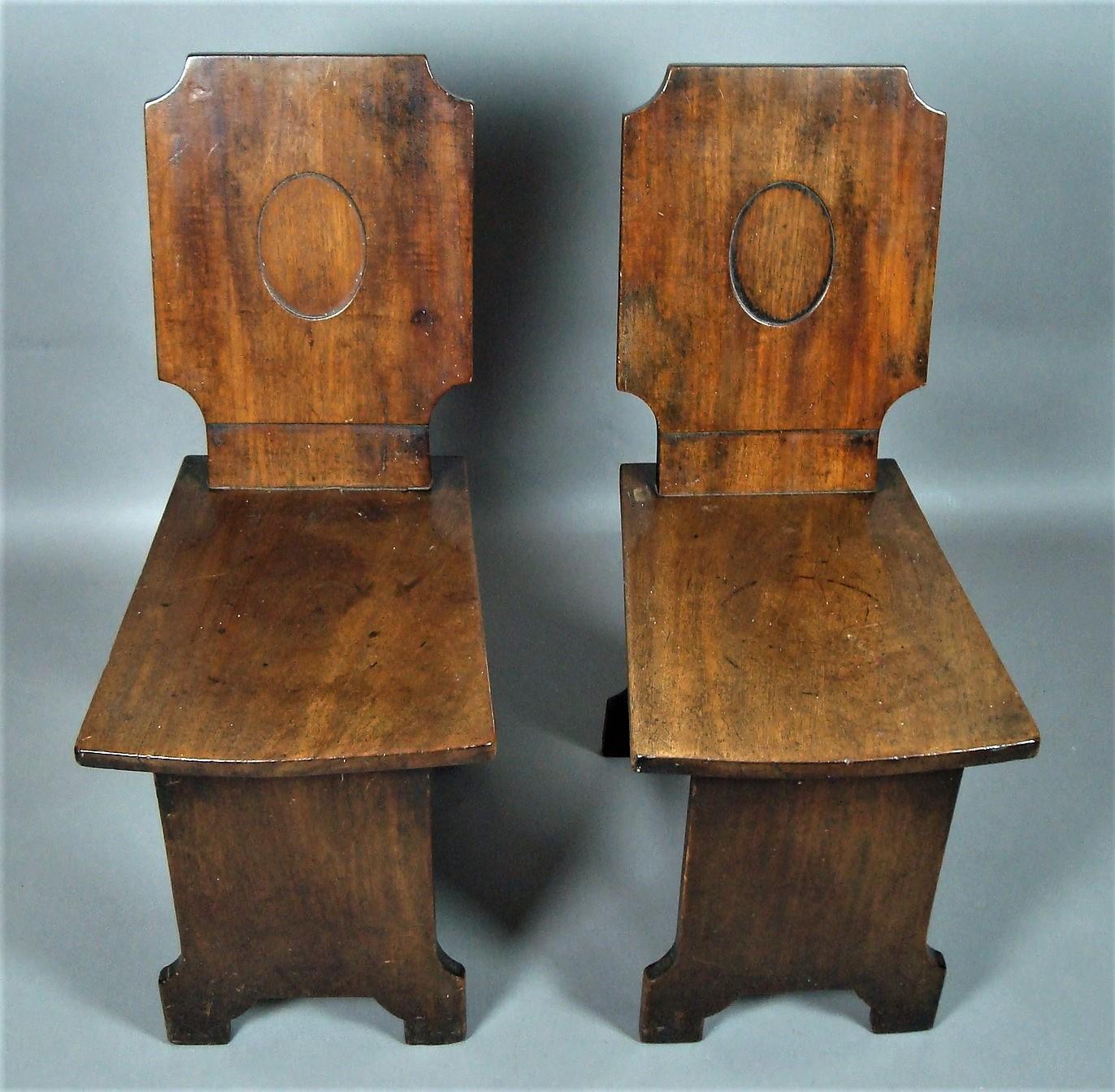 Regency pair of mahogany hall chairs, of very unusual restrained design; the tablet shaped backs incorporating dished oval panels, above a simple tapering seat of dished form, raised on slab supports with shaped feet united by a simple