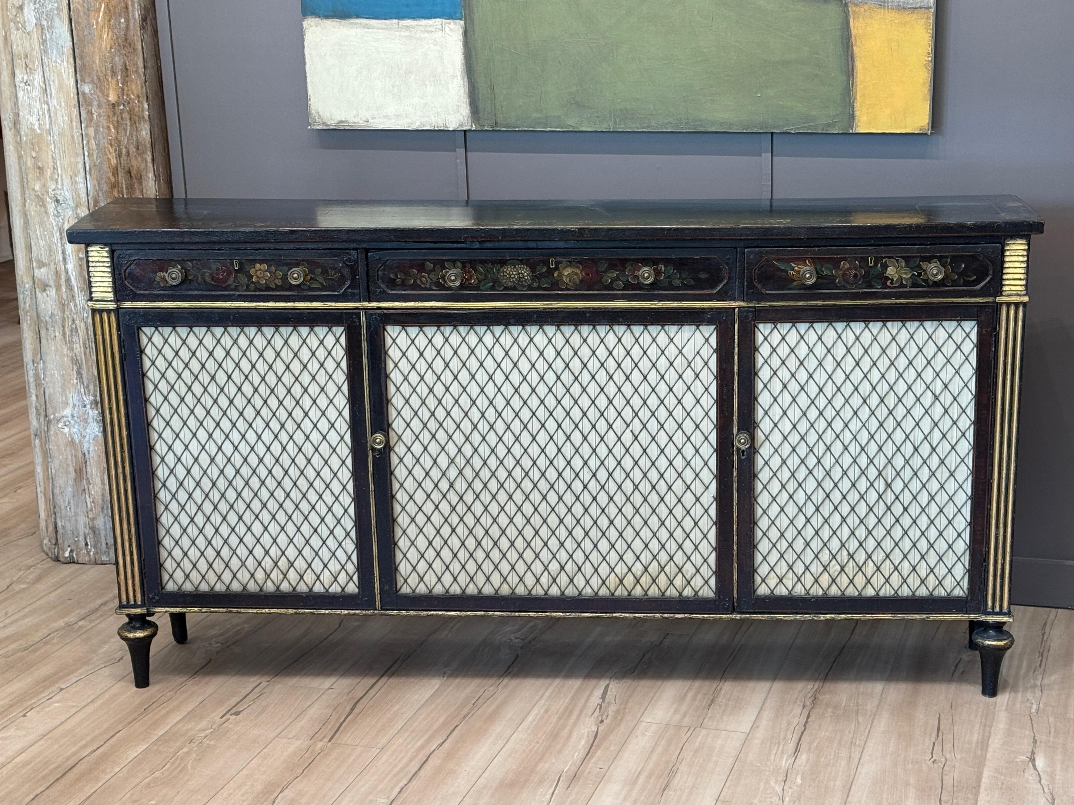 An exceptional Regency period cabinet, parcel-gilt and ebonized, with hand-painted floral decoration across three drawers. The lower doors feature original inset silk panels protected by a diamond-patterned wire grille. Fluted corner stiles and