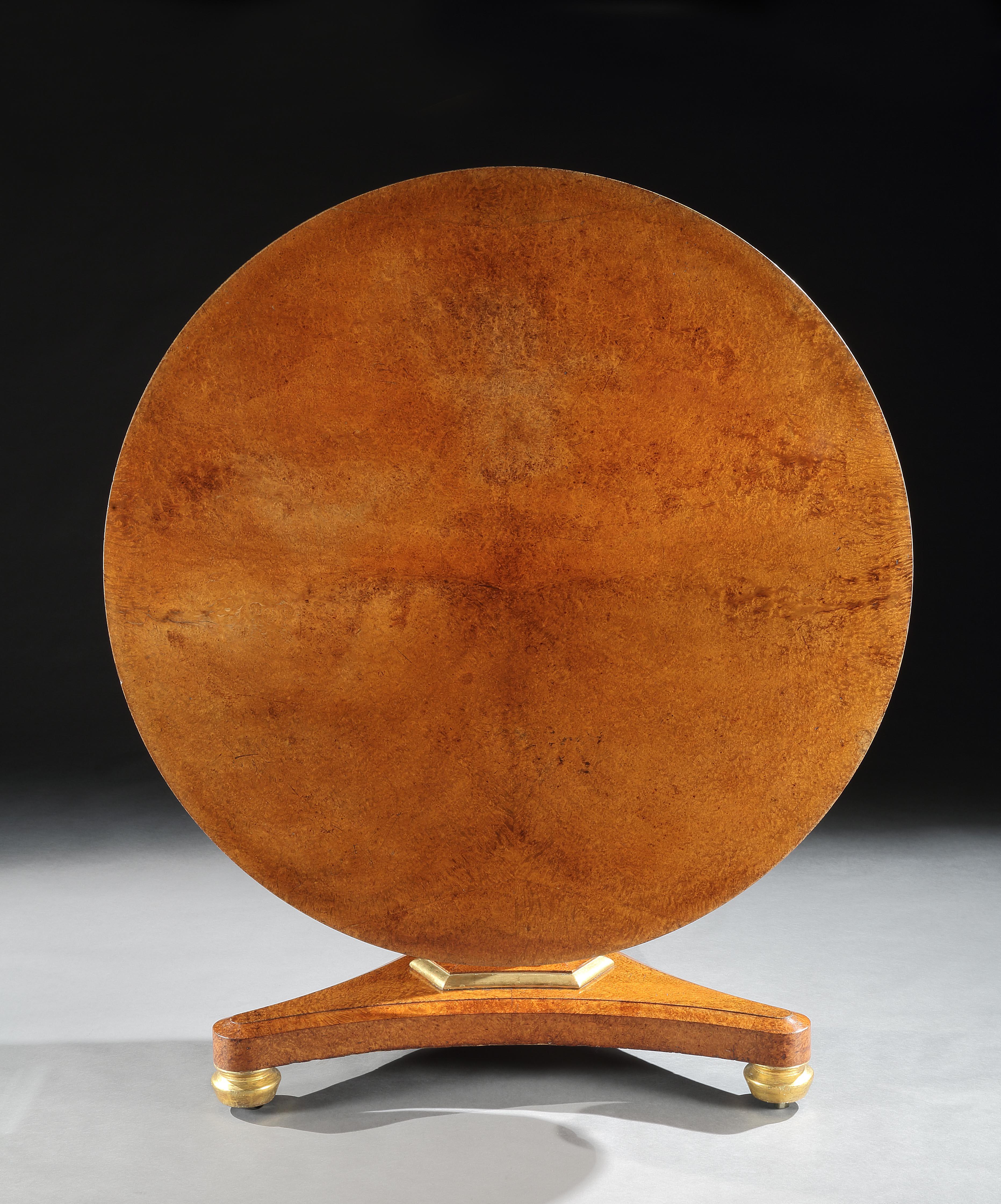 The circular top veneered in finely figured amboyna retaining a superb colour and patina, above a slim frieze with a gilded line, the table supported on a triangular stem and base similarly veneered in amboyna, and with gilded mouldings and standing