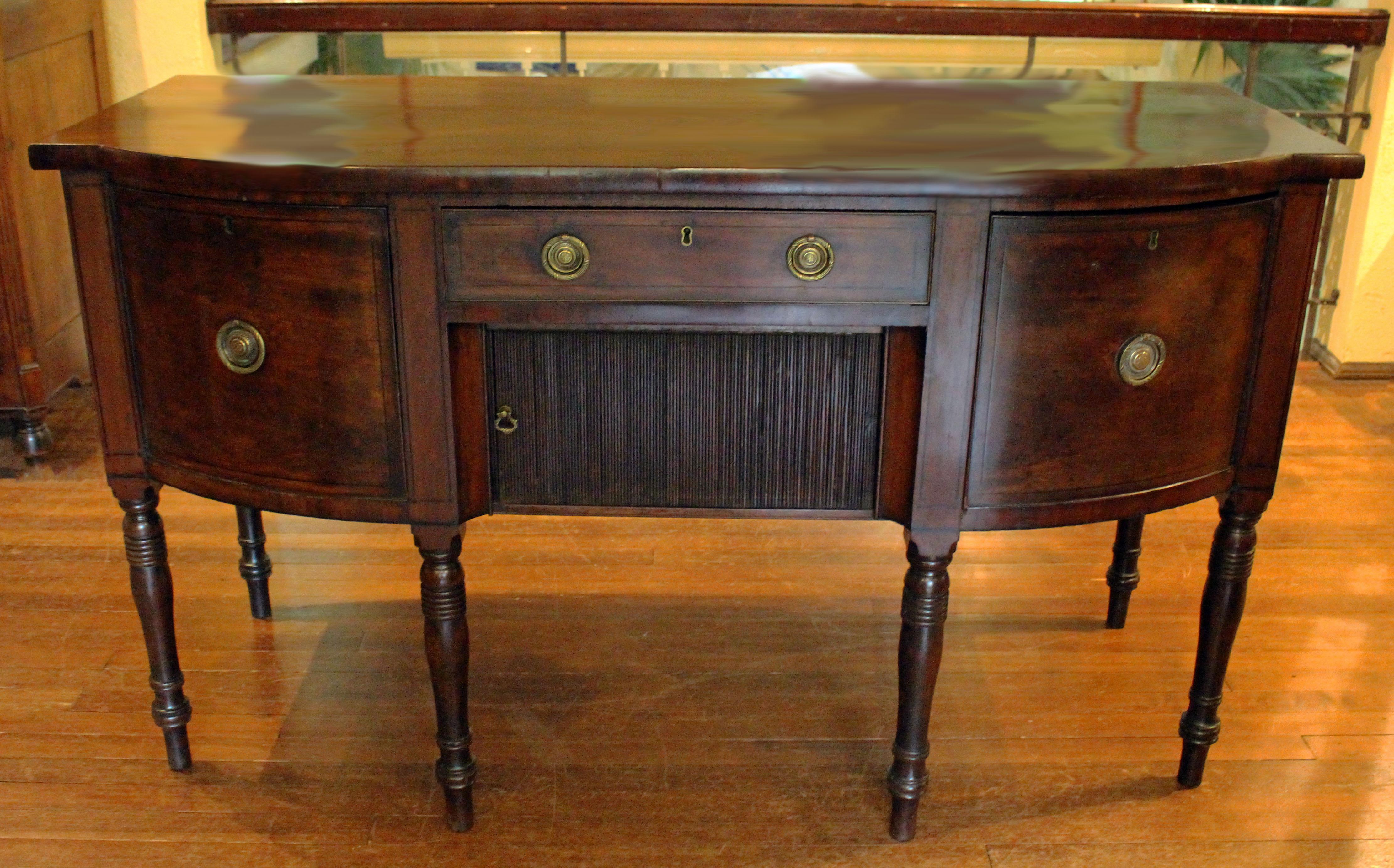 A bowfront sideboard, c. 1815, Regency period in the Sheraton taste. Raised on 6 turned, tapered legs with ring turned accents. Central drawer over recessed tambour door cabinet, flanked by a cellarette drawer and a cabinet. Ebony line & dot inlays.