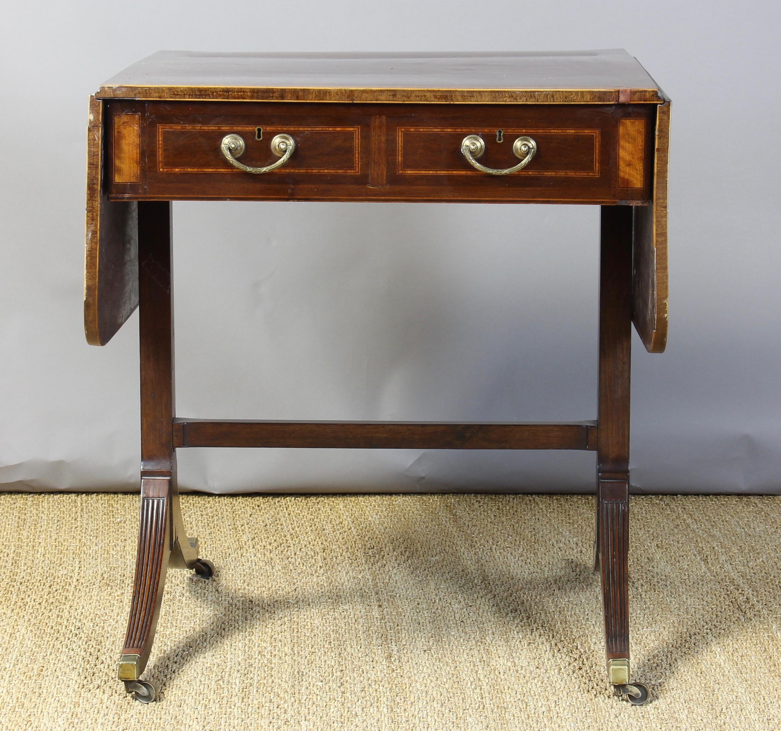 An early 19th century English mahogany drop-leaf sofa table with satinwood inlay and original brass hardware on two working drawers and two dummy drawers resting on reeded saber legs terminating in brass casters.