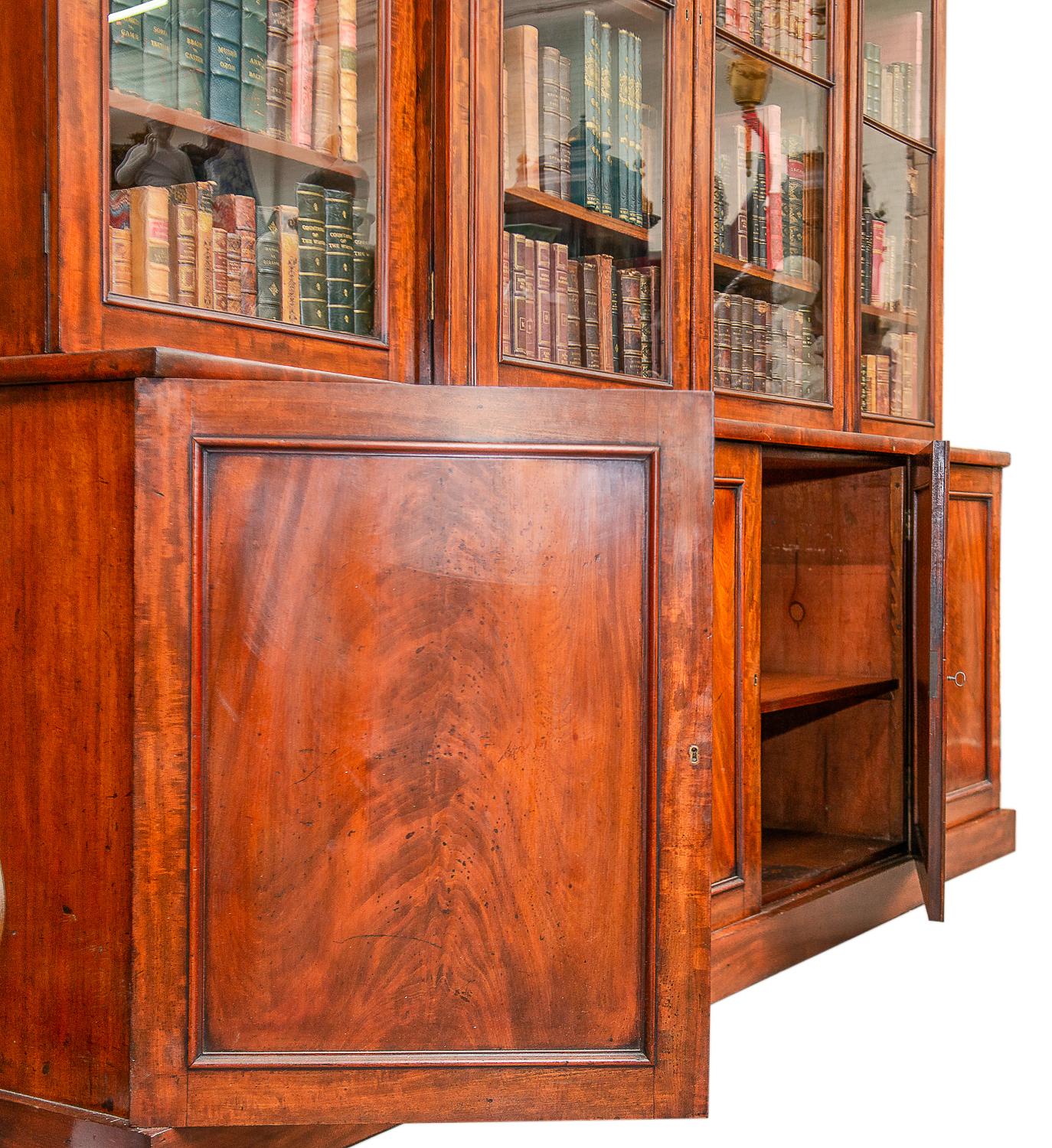 A very good quality Regency period mahogany breakfronted library bookcase, having four glazed doors above with adjustable shelves within. Inset molded panels with roundels above. Four paneled doors beneath with figured mahogany veneers and raised on