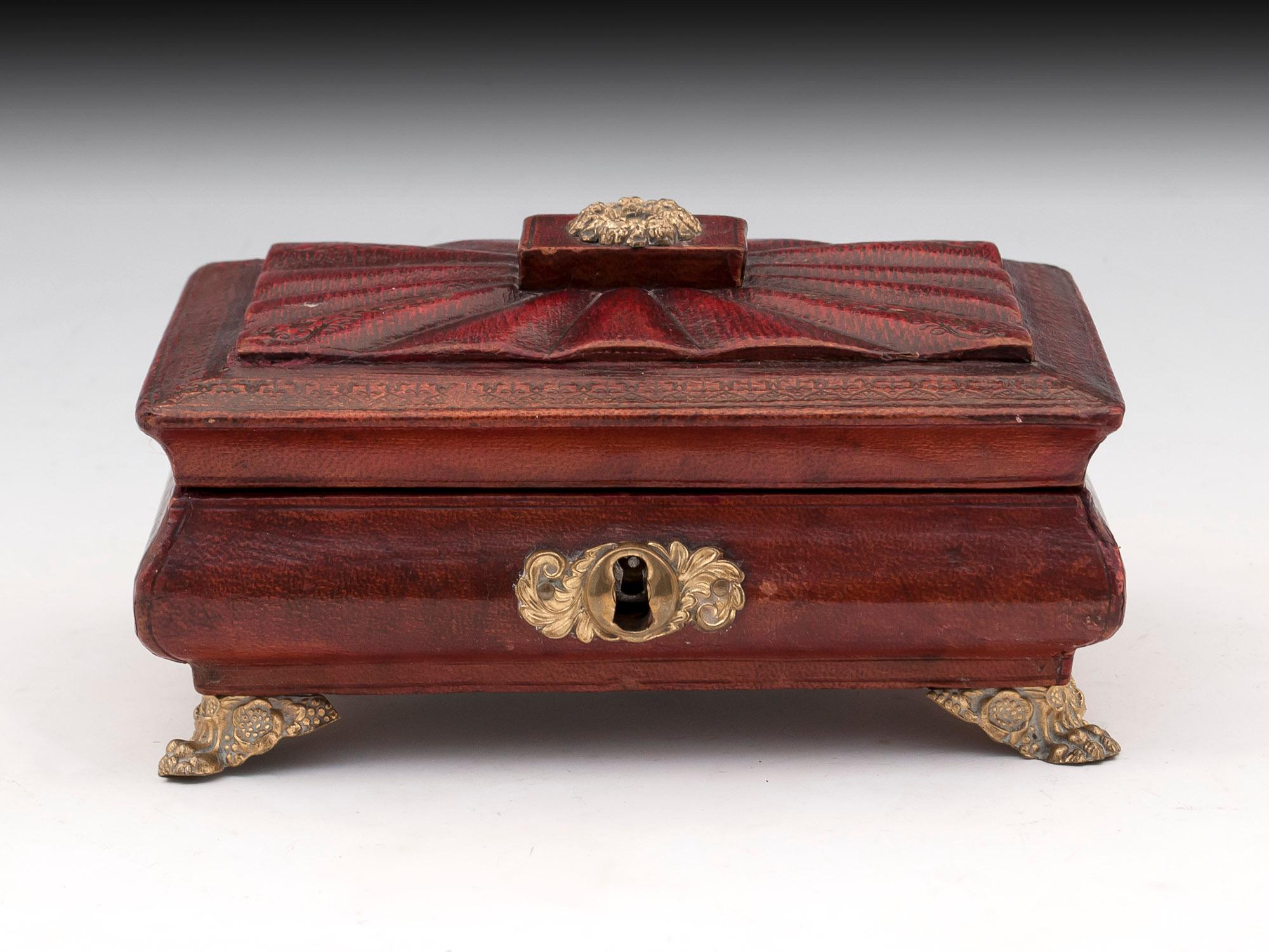 Antique red leather sewing box with ornate brass escutcheon and bracket feet. 
The blue paper lined interior features various sewing supplies including six thread reels, Bone pricker, needle case, brass thimble, and a pincushion.