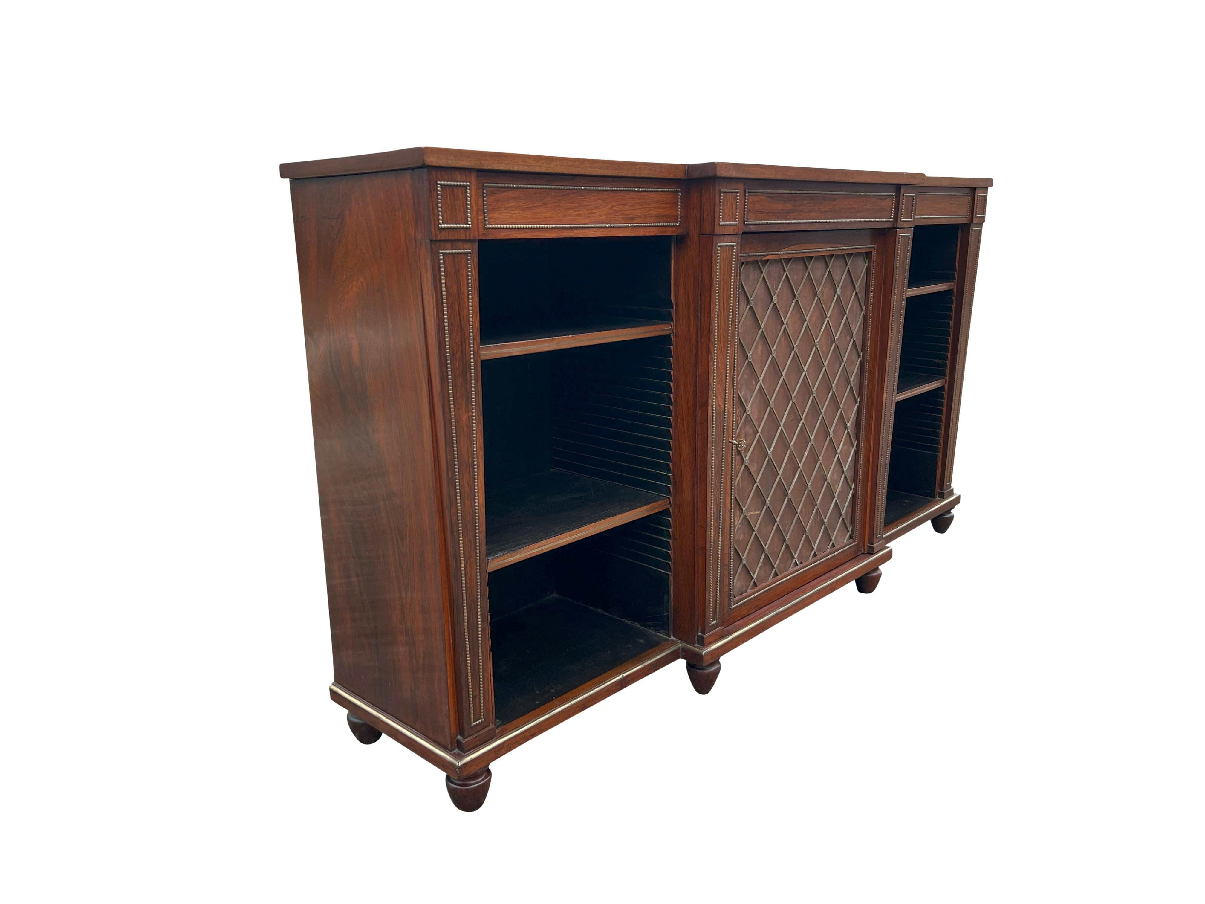 With a rectangular breakfront form top over a central door with brass grillwork. The silk under brass needs replacement or can be removed and just have open brass work. Flanked by two open cases with two shelves for each. All surrounded by brass