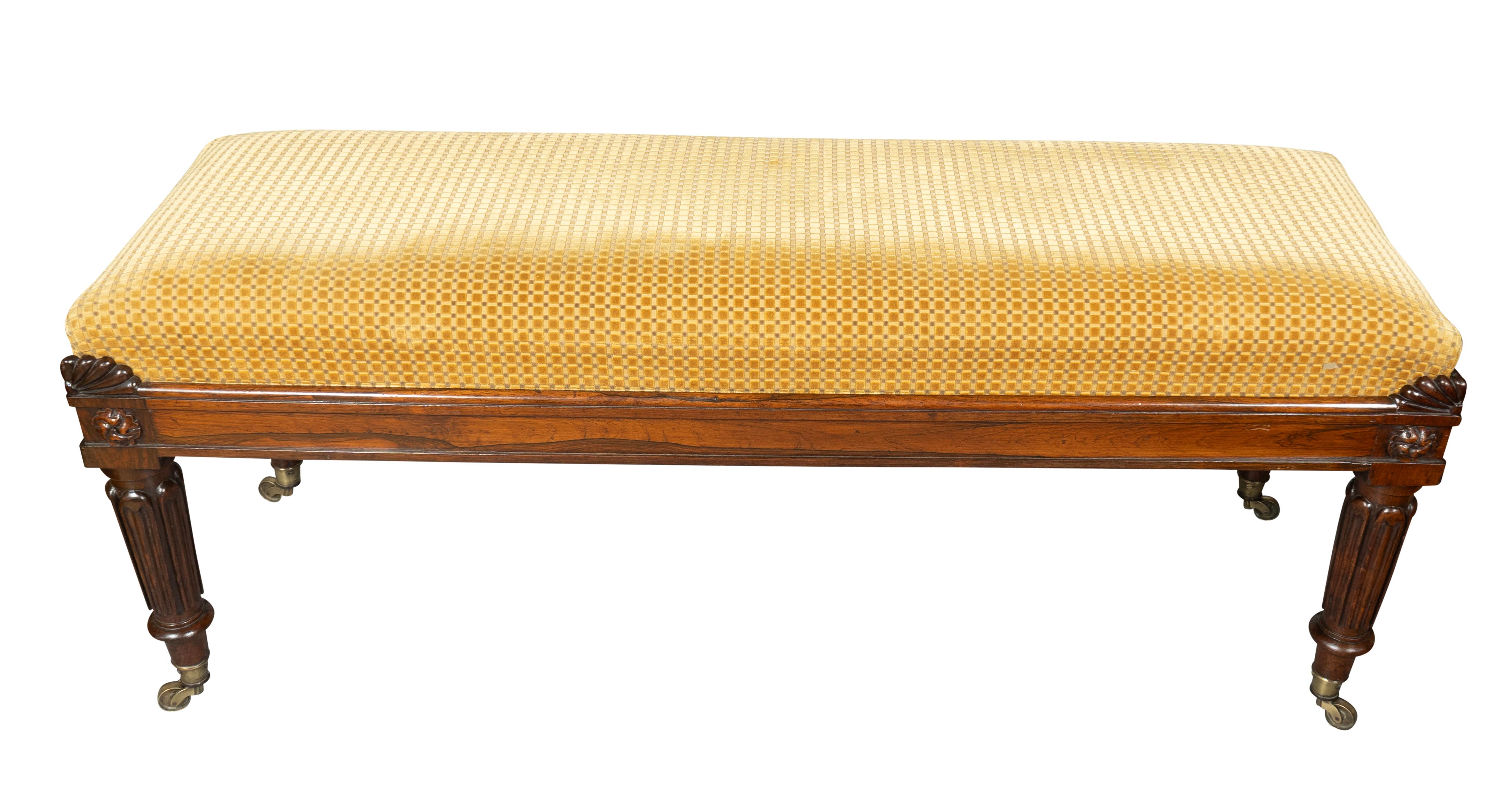 Asiento rectangular tapizado. Las esquinas con talla estriada. La barandilla del asiento con laterales moldeados y rosetas talladas. Patas circulares ahusadas con lengüeta. Ruedas para tazas.