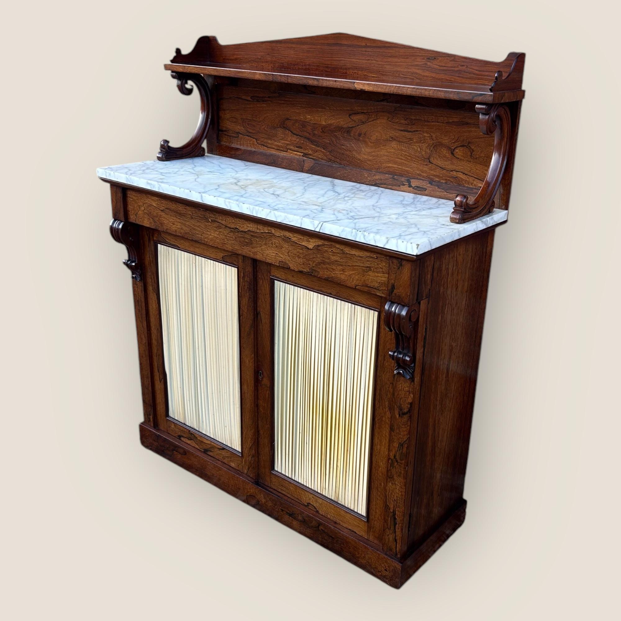Qui abbiamo un Chiffoniere Regency in legno di palissandro con un piano in marmo bianco e grigio. In cima al chiffonier c'è un'unica mensola a gallerie sostenuta da supporti a cigno con volute. C'è un cassetto con fregio che si apre su un interno a