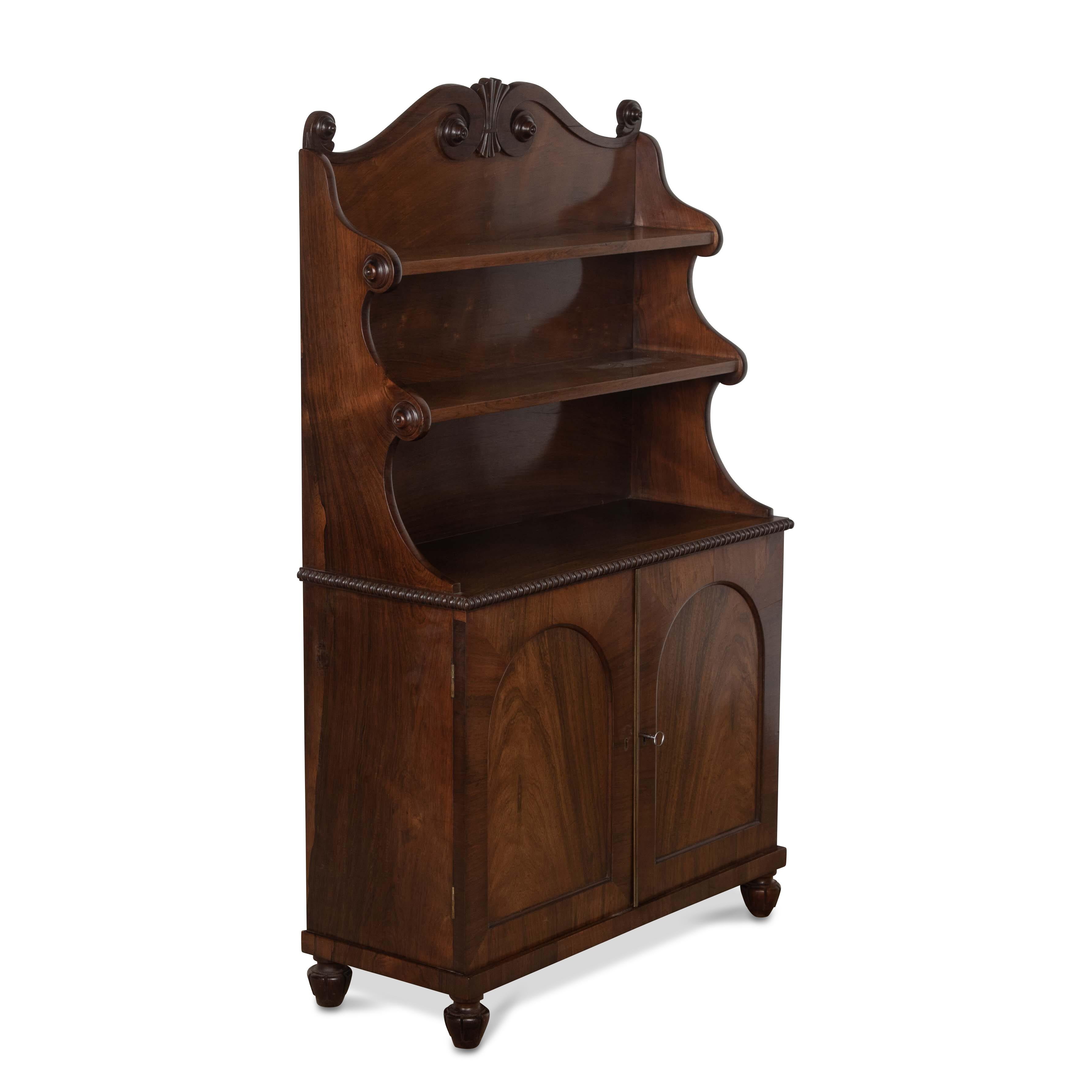 A regency rosewood side cabinet/bookcase of small proportions, the waterfall superstructure and scroll pediment with roundels. Above a cupboard base with a pair of arched panel doors opening to an adjustable shelf interior. Raised on tulip carved