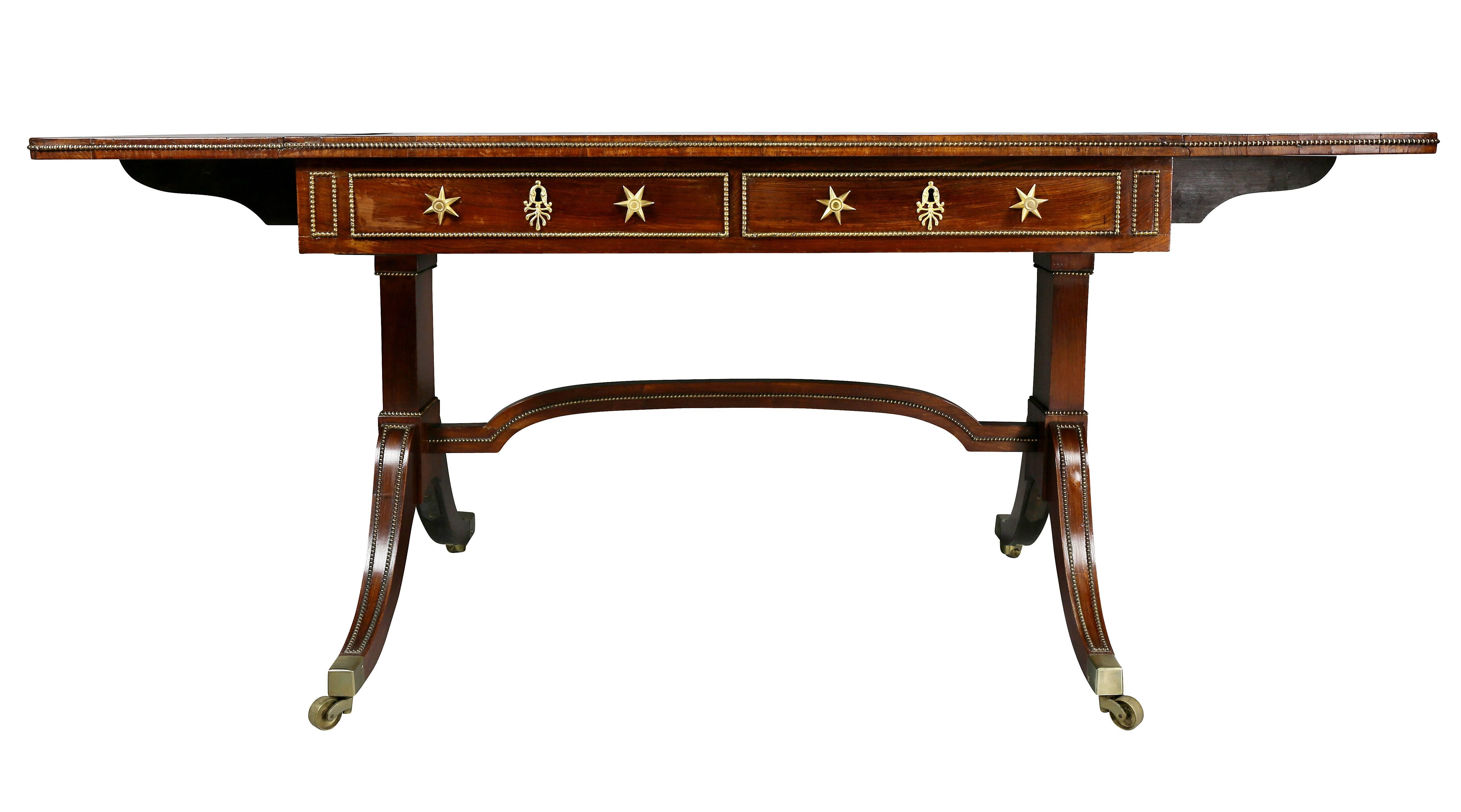 With rectangular drop leaves and beaded edge over two drawers with star form handle backplates over a trestle support joined by a stretcher raised on saber legs with casters.