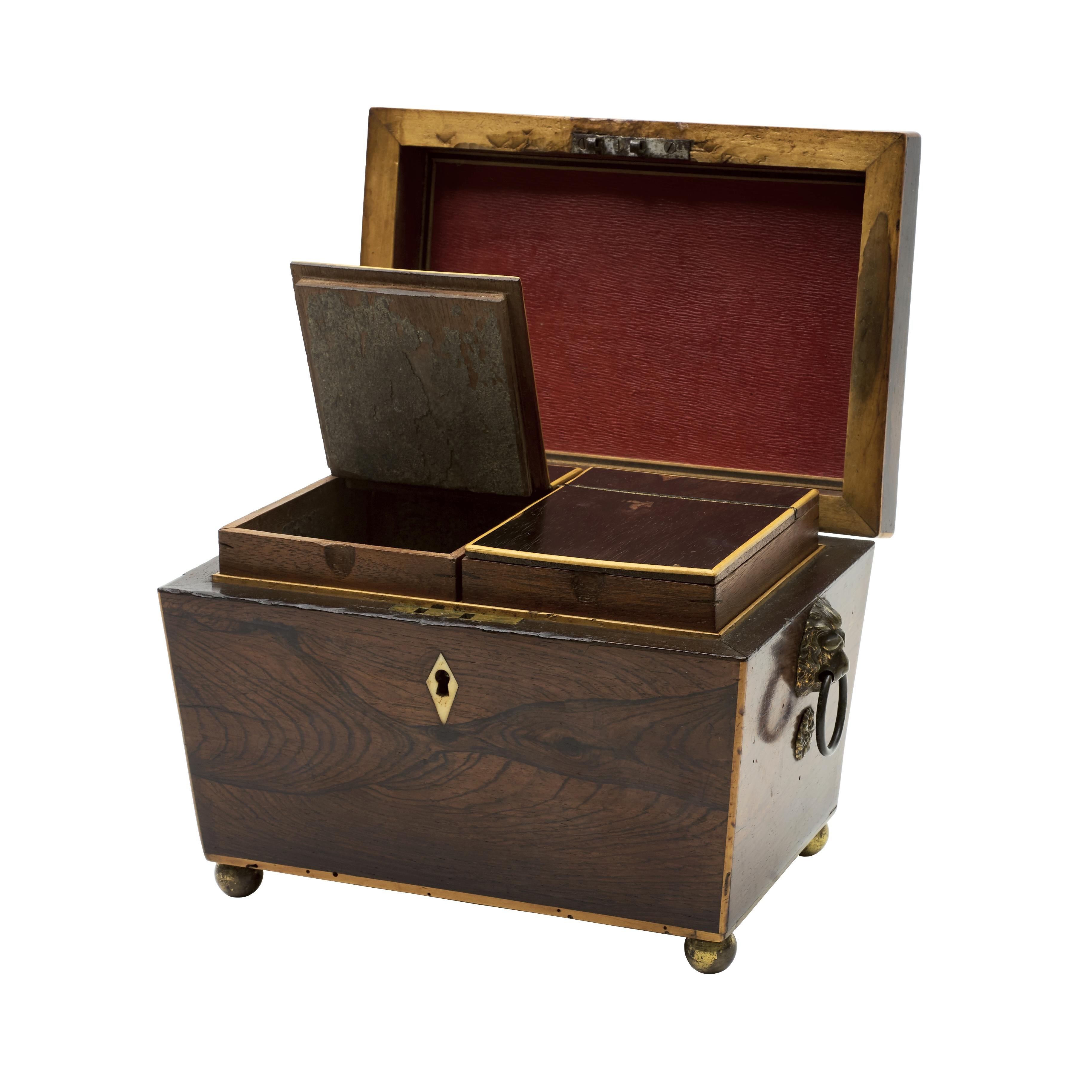 A rosewood pedestal tea caddy, made in England during the Regency period.
The caddy is veneered with rosewood and banded with satinwood. There are gilt-brass handles (lion masks) on either side as well as four spherical feet to the base. The box