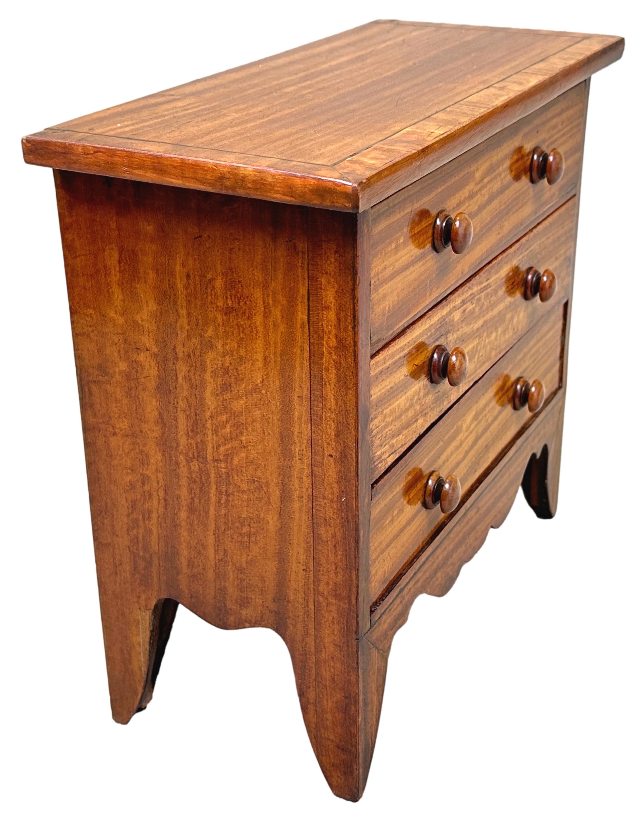 Une commode miniature en bois satiné de bonne qualité, de la fin de la période Régence, 19ème siècle, de petites proportions attrayantes, avec un dessus bien figuré et à bandes croisées, au-dessus de trois tiroirs avec des boutons en bois tournés