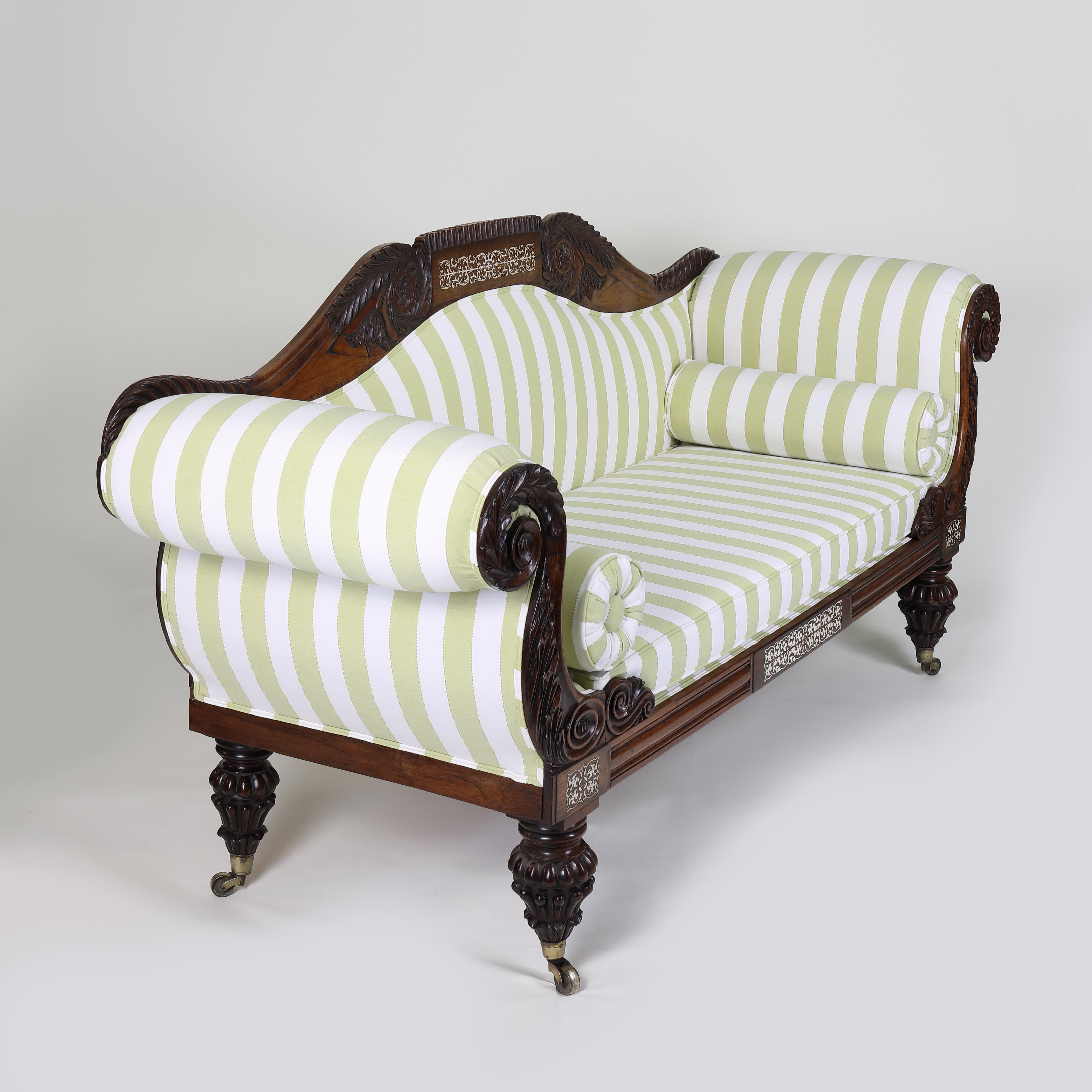 A grand Regency scroll-end settee constructed in rosewood with panels of intricately cut ivory. Adorned with acanthus leaf carving to the back and scroll ends and standing on turned and reeded legs with brass castors. 
An opulent and imposing piece