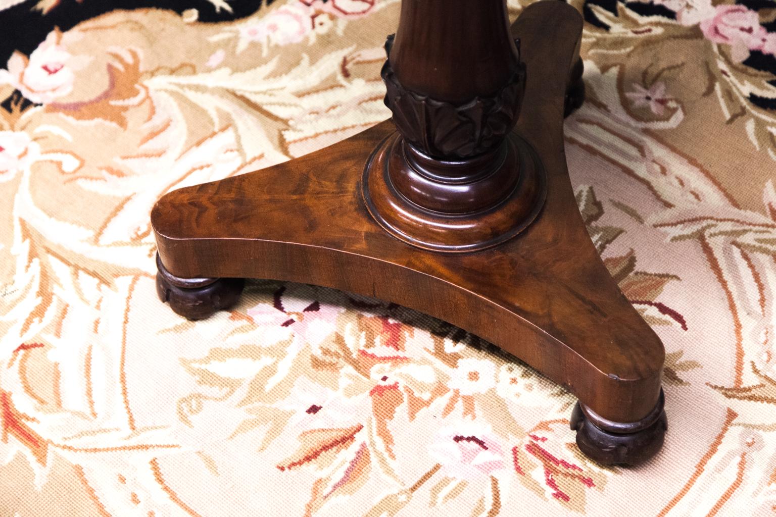This Regency sewing table is mahogany with hinged lift top. The body is faceted with shaped apron and acanthus carved stem on a trifid shaped platform base. It rests on carved bun feet.