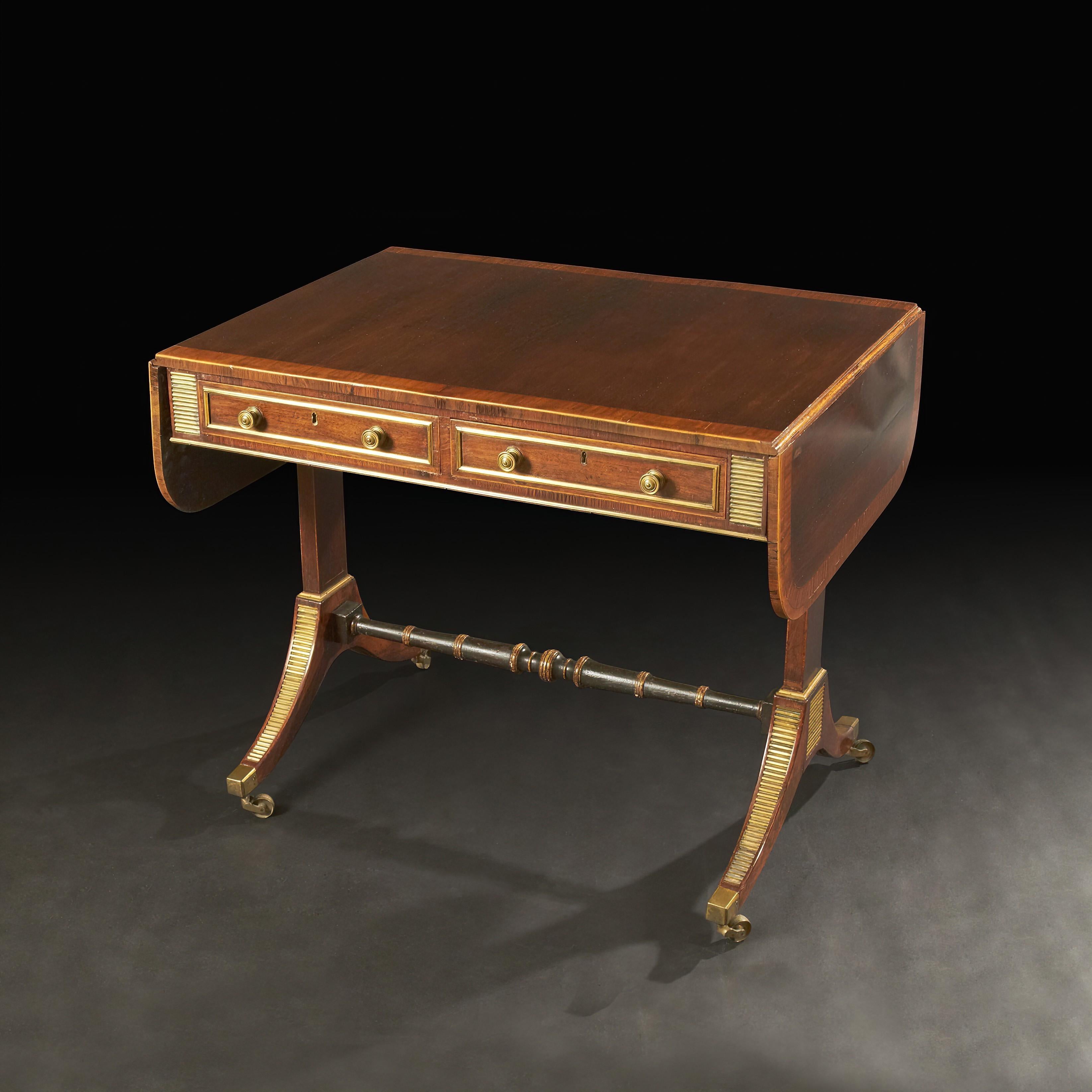 A fine English Regency rosewood and satinwood cross-banded sofa table, with D form drop leaf ends, two drawers with brass hardware and trim, with a turned ebonized and gilded stretcher base, with brass mounts and raised and brass casters. Attributed