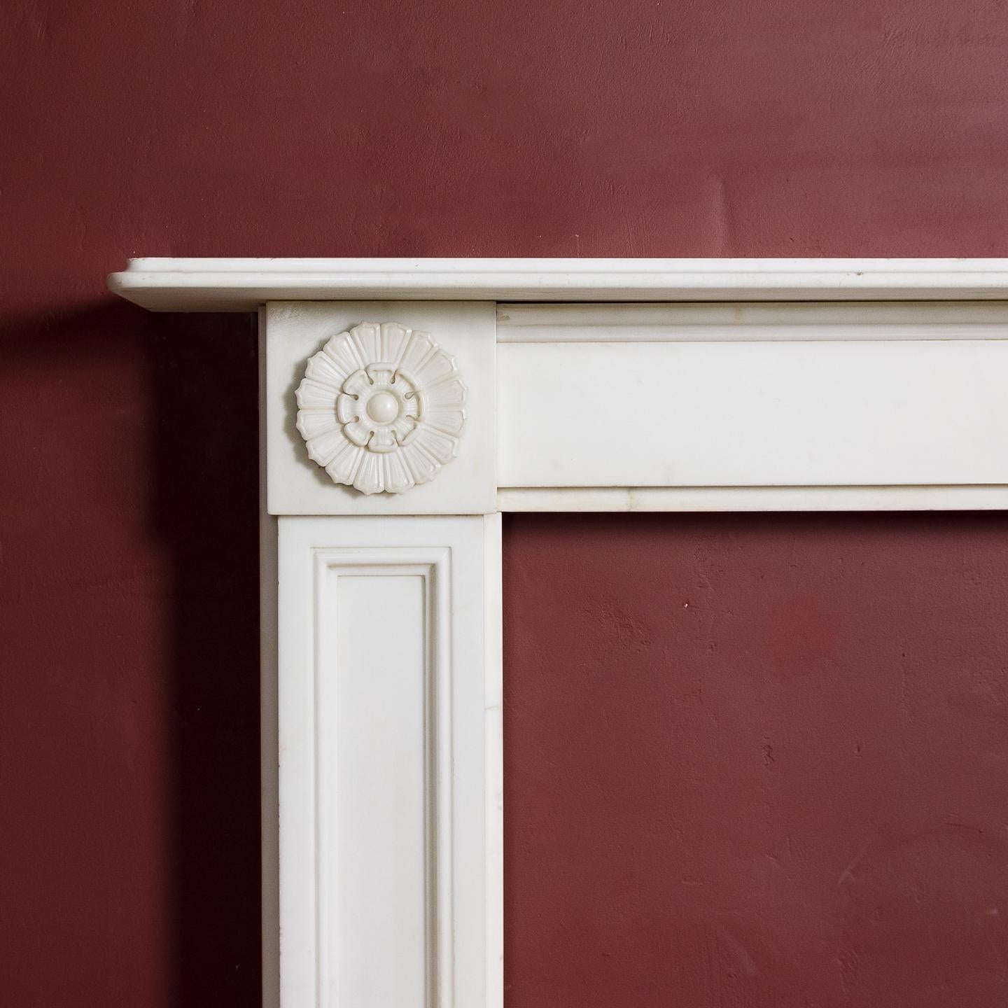 Regency statuary marble fireplace, in the manner of Sir John Soane, the thumb moulded shelf above plain frieze flanked by flower head corner blocks, on panelled jambs.

  