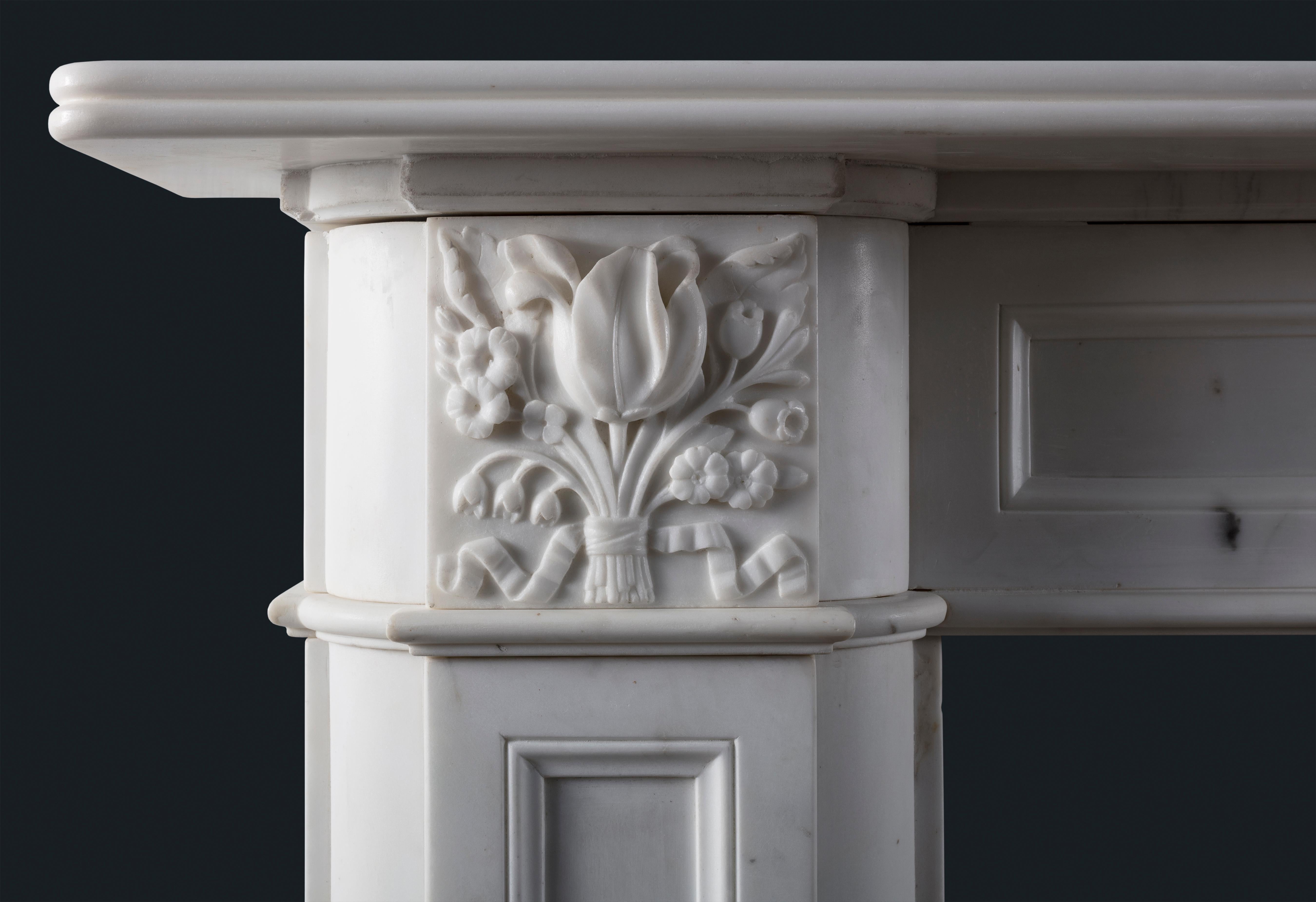 An early-to-mid nineteenth century Regency period fireplace surround in Statuary marble, the shelf with single reed above fielded panel frieze and jambs, the corner-blockings depicting bunched flowers, all raised on block feet.

English,