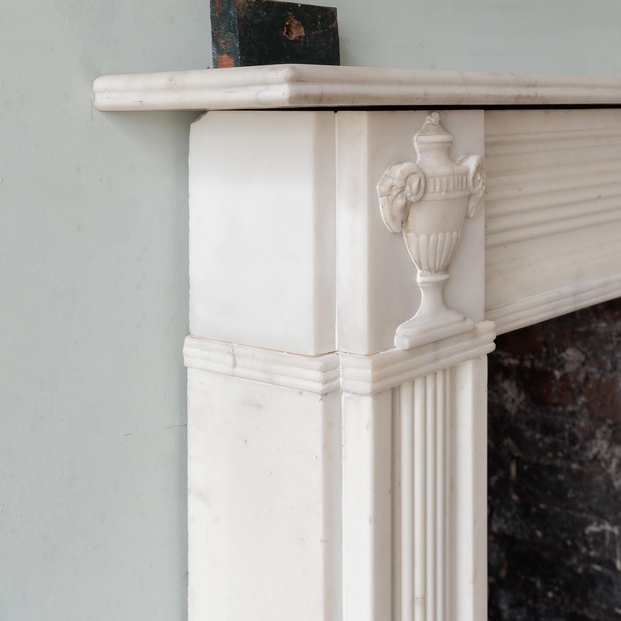 A Regency statuary marble neo-classical fireplace, the rectangular shelf with single reed above frieze centred by tablet with a shallow lobed urn with ram's heads to either side, the carved corner blocks showing conforming classical urns, the frieze