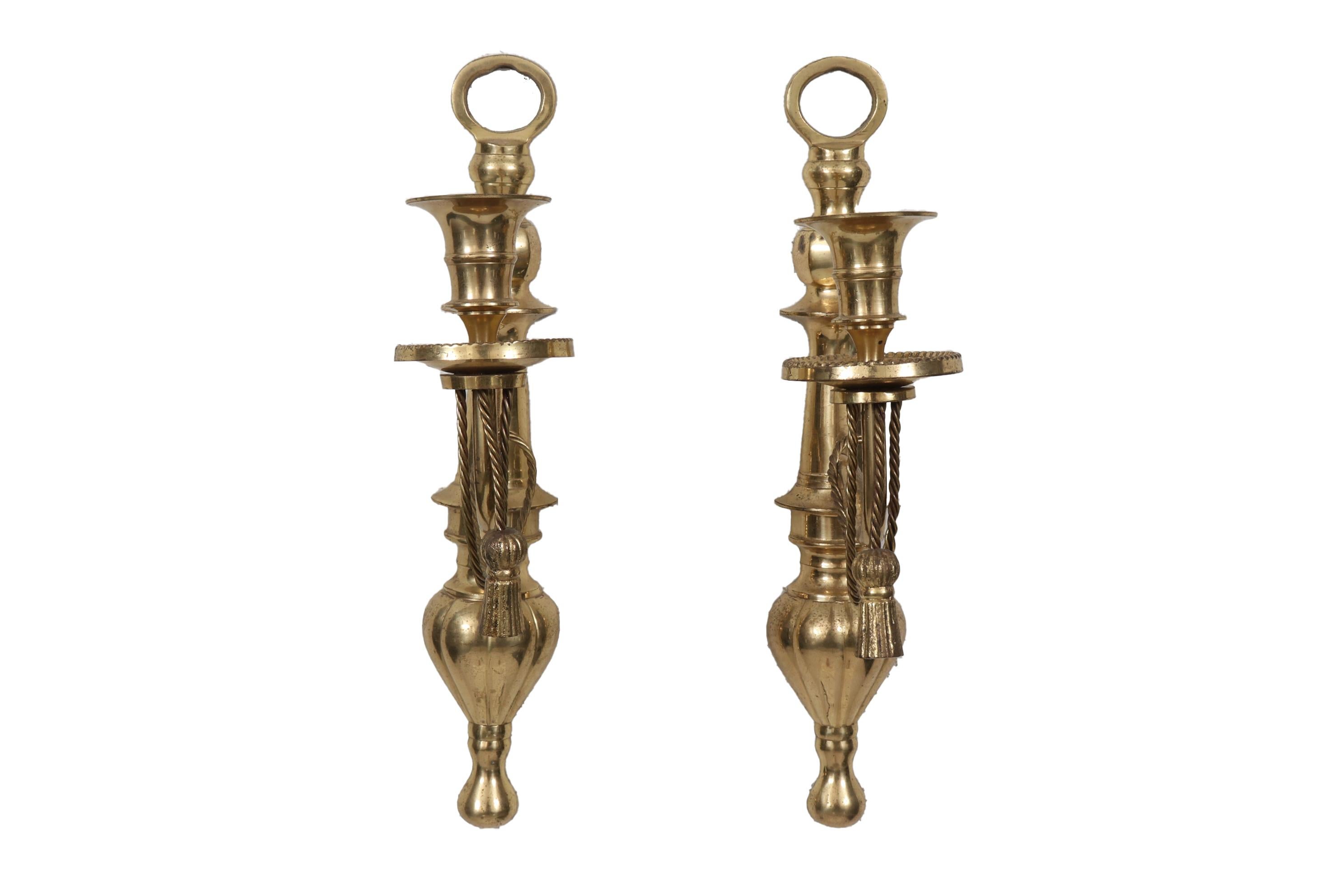 A pair of Regency style brass candlestick sconces. Slender curved arms that support a single capital are decorated with brass tassels and rope. Supported on turned baluster shaped back plates. Dimensions per sconce.