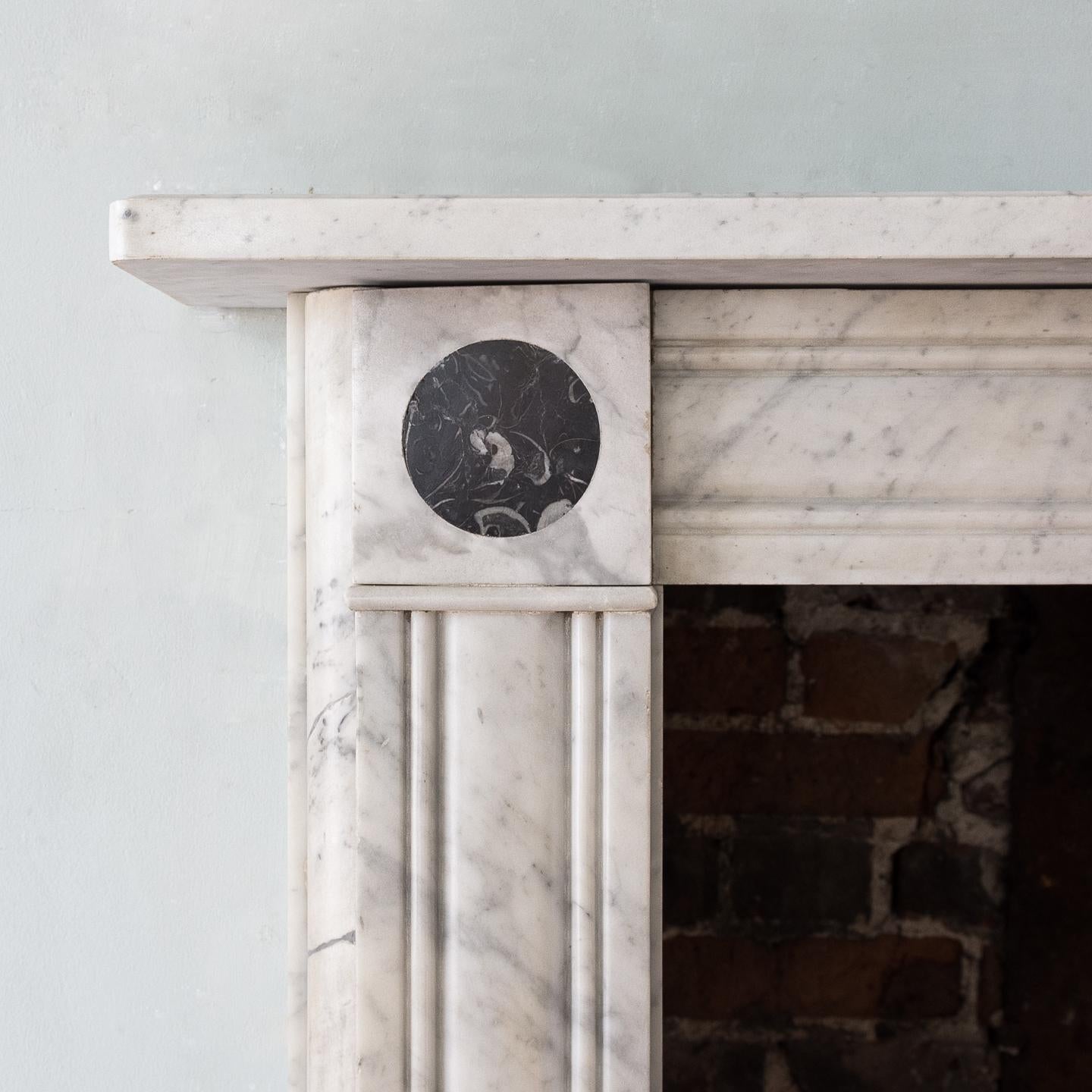 Regency style Carrara marble fireplace, the plain rectangular shelf above moulded frieze flanked by Peacock's eye marble roundels, the jambs with conforming moulded detail, on square foot blocks, 20th century.

Measures: Opening width 92.5 cm x