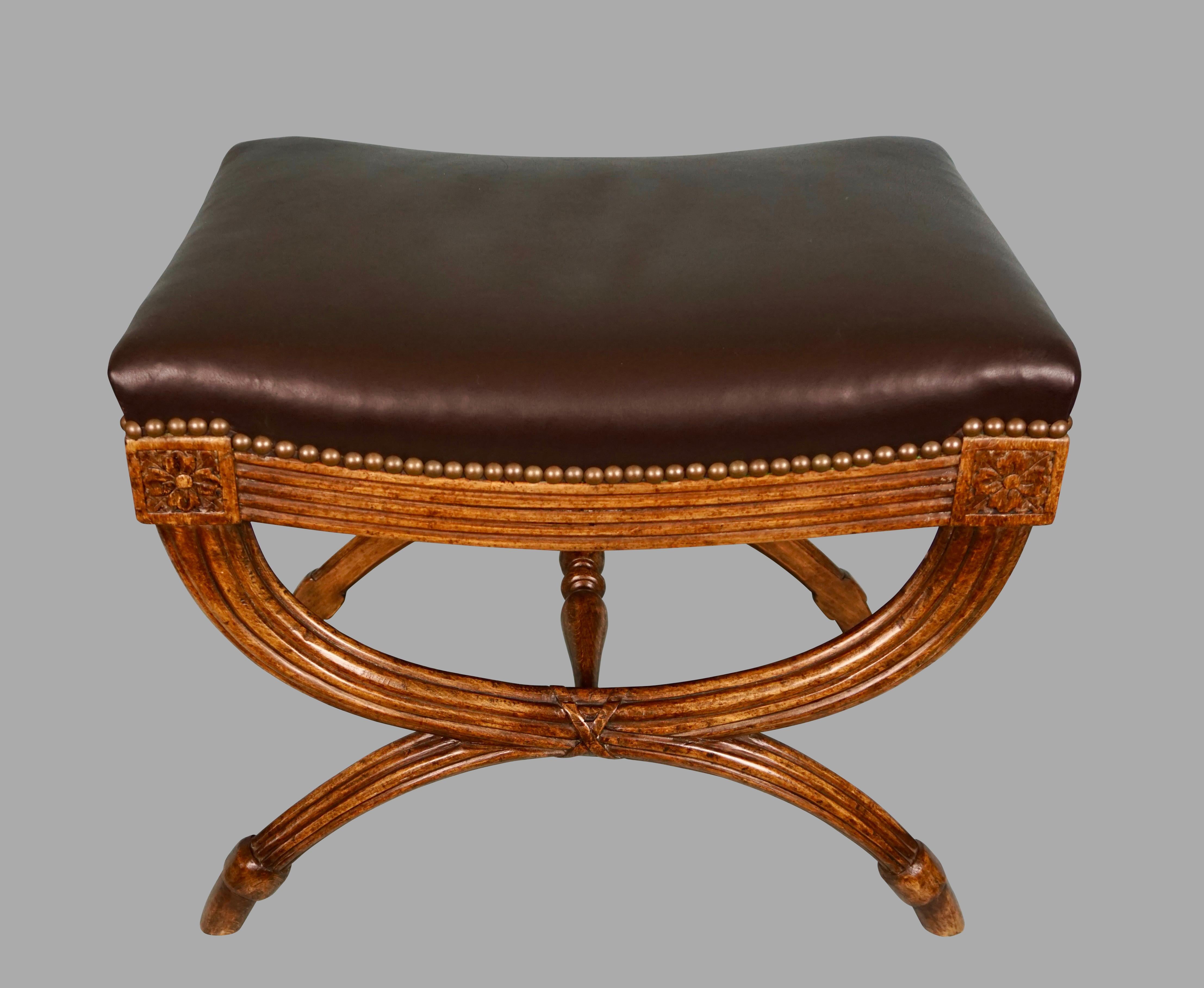An elegant English Regency style mahogany curule form bench, the reeded frame terminating in carved floral bosses, the legs connected by a turned stretcher and ending in hoof feet. The piece is handsomely upholstered in black hide with brass