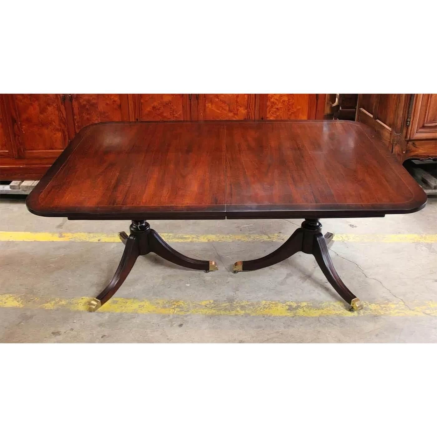 An English Regency style mahogany double pedestal extending dining table with a cross banded and line inlaid edge, four 12 inch leaves stored inside the table and raised on two-column pillar three-leg pedestals raised on brass castors. By Kindel,