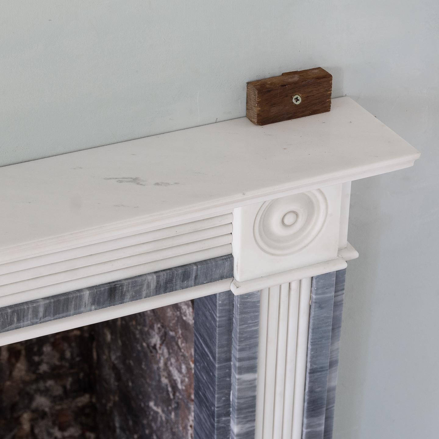 A Regency style dove grey and statuary marble chimneypiece, circa 1900, the moulded shelf above reeded frieze centred by carved tablet, the bullseye end-blocks above conforming reeded jambs, on plain foot blocks.
  