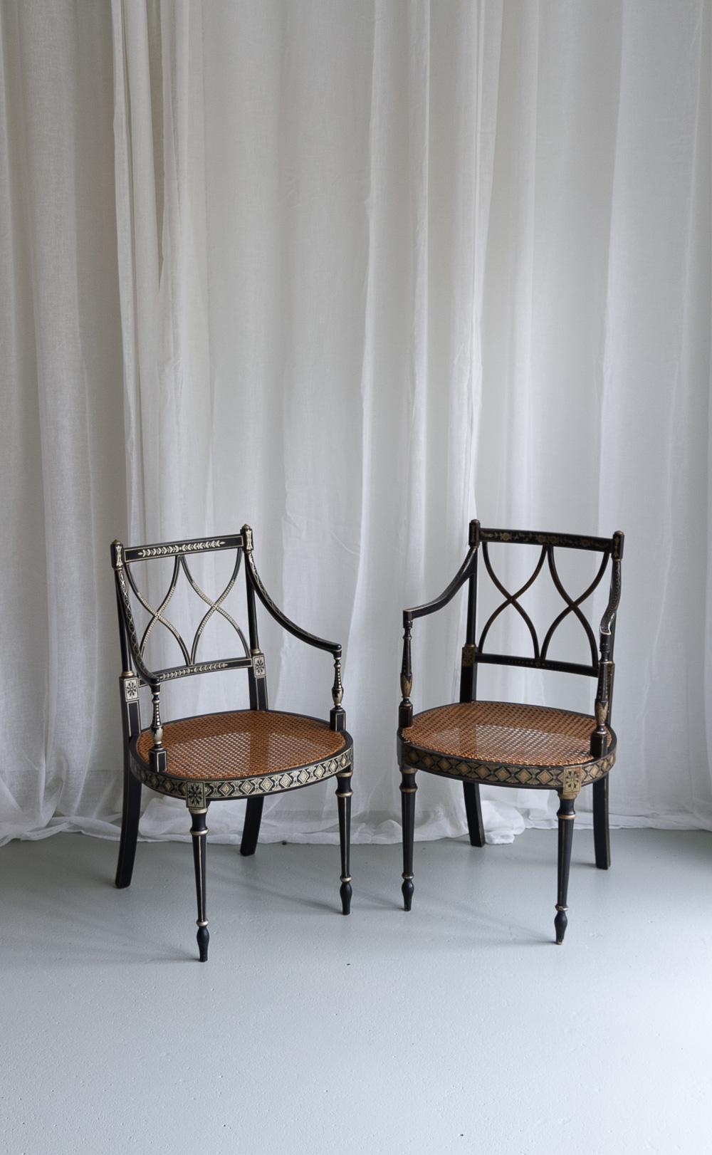 Regency Style Ebonized Cane Armchairs, Set of 2.
Pair of English Regency style black lacquered chairs with caned seat and hand painted gold motifs circa 1970s.
One chair has a bit more patina and a handwoven seat and therefore may be a bit
