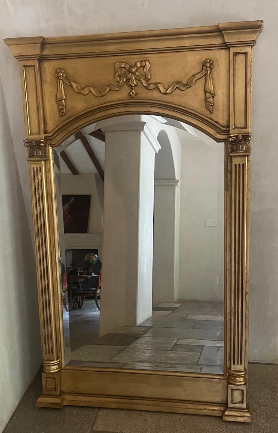 Miroir biseauté vintage du 20ème siècle de style Régence. De grande taille, la partie supérieure est une plaque rectangulaire surmontée de guirlandes sculptées et nouées par des rubans et flanquée de colonnes cannelées corinthiennes.

Peut être