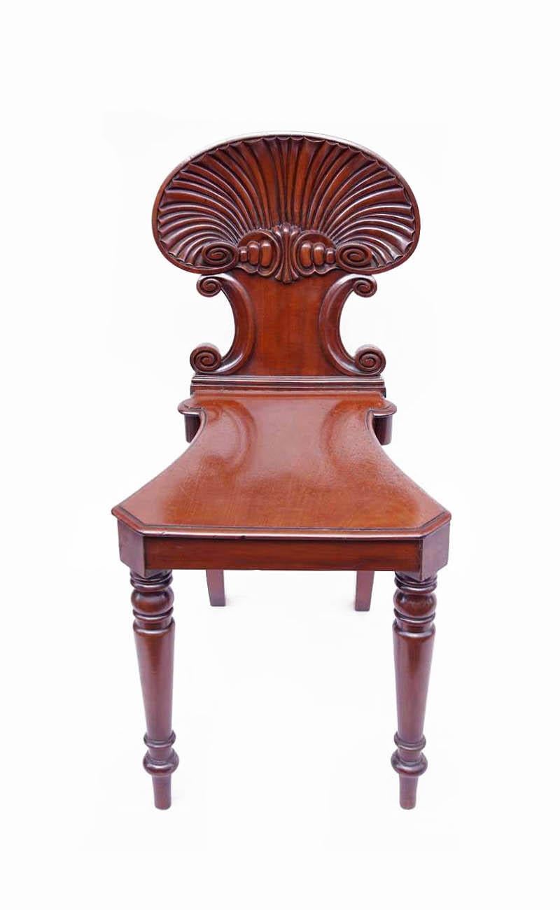 Regency style carved mahogany hall chair, standing on two turned tapered front legs and two saber back legs. Seat with canted corners tightened backwards. Back carved as shells with two C-scrolls beneath.
Model created by Gillows of London and
