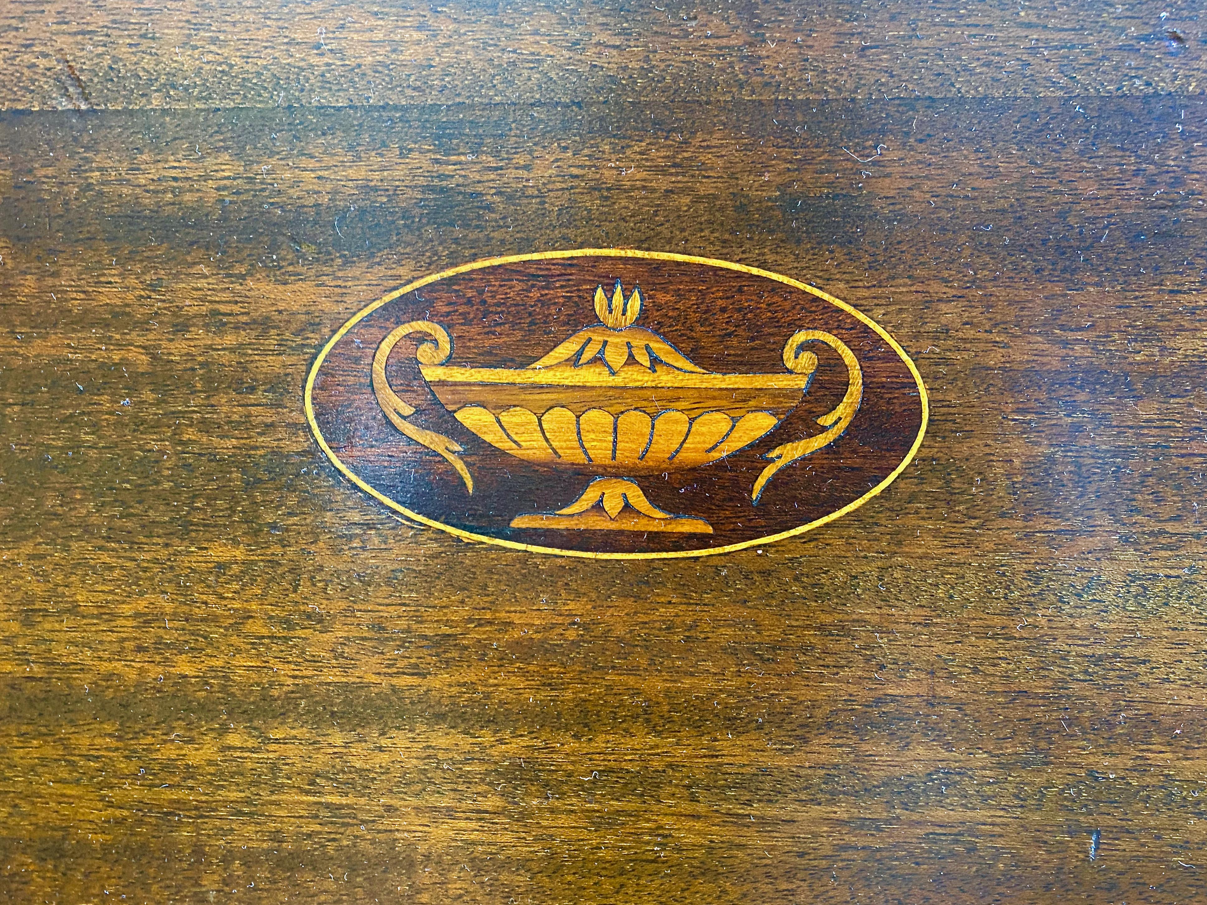 This is a good example of a George III-style Victorian Oval Butler's Tray that dates to c. 1885-1890. The fine mahogany tray is detailed with an inlaid satinwood neoclassical-style lidded urn and brass side handles. The tray is in overall very good