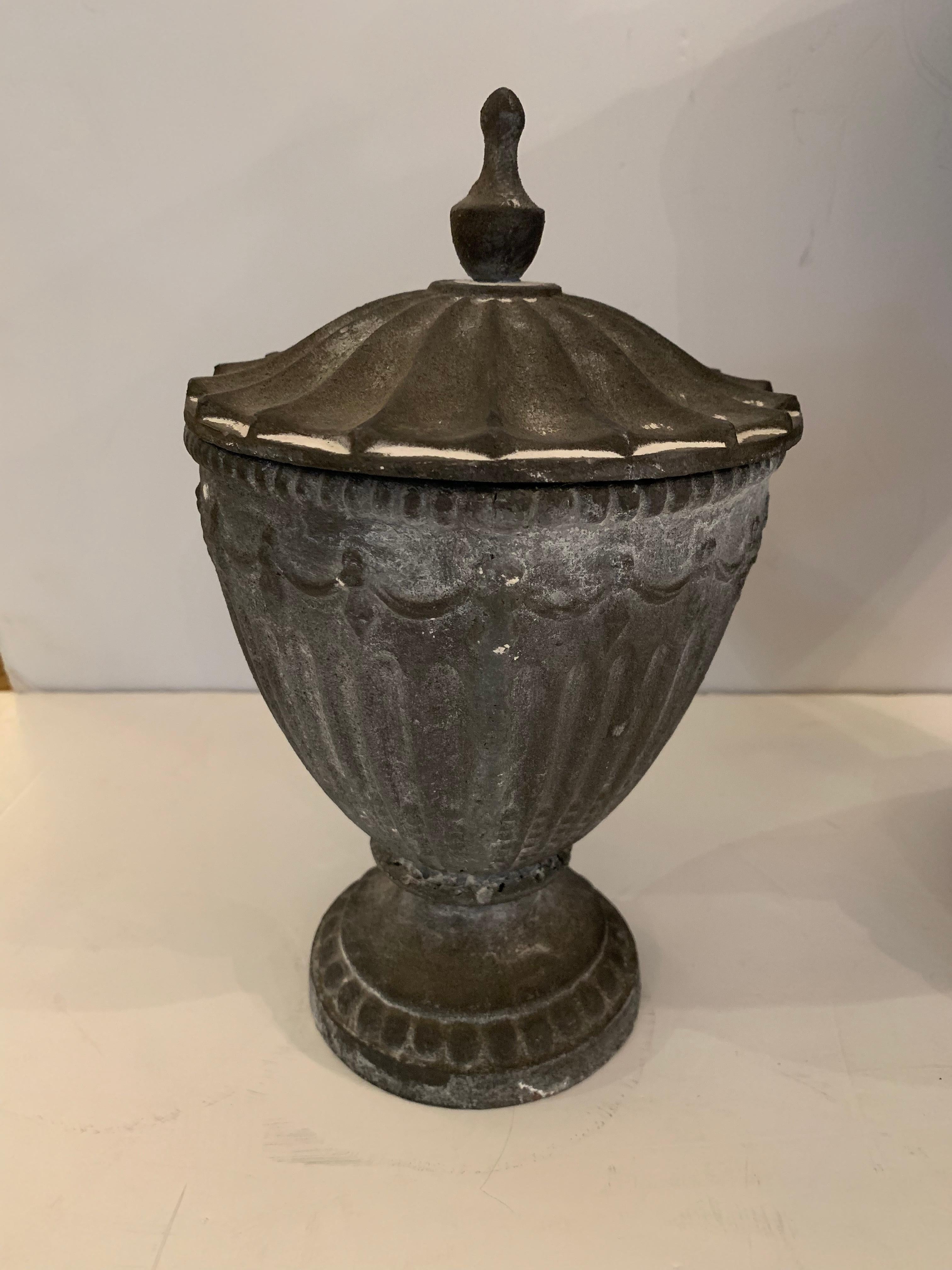 A lovely pair of oramental classical jardinières having fluted bodies in two sizes, resting on round bases, complete with faceted lids, swag moldings, and original finials. The naturally patinated cast iron has remnants of white paint, and is