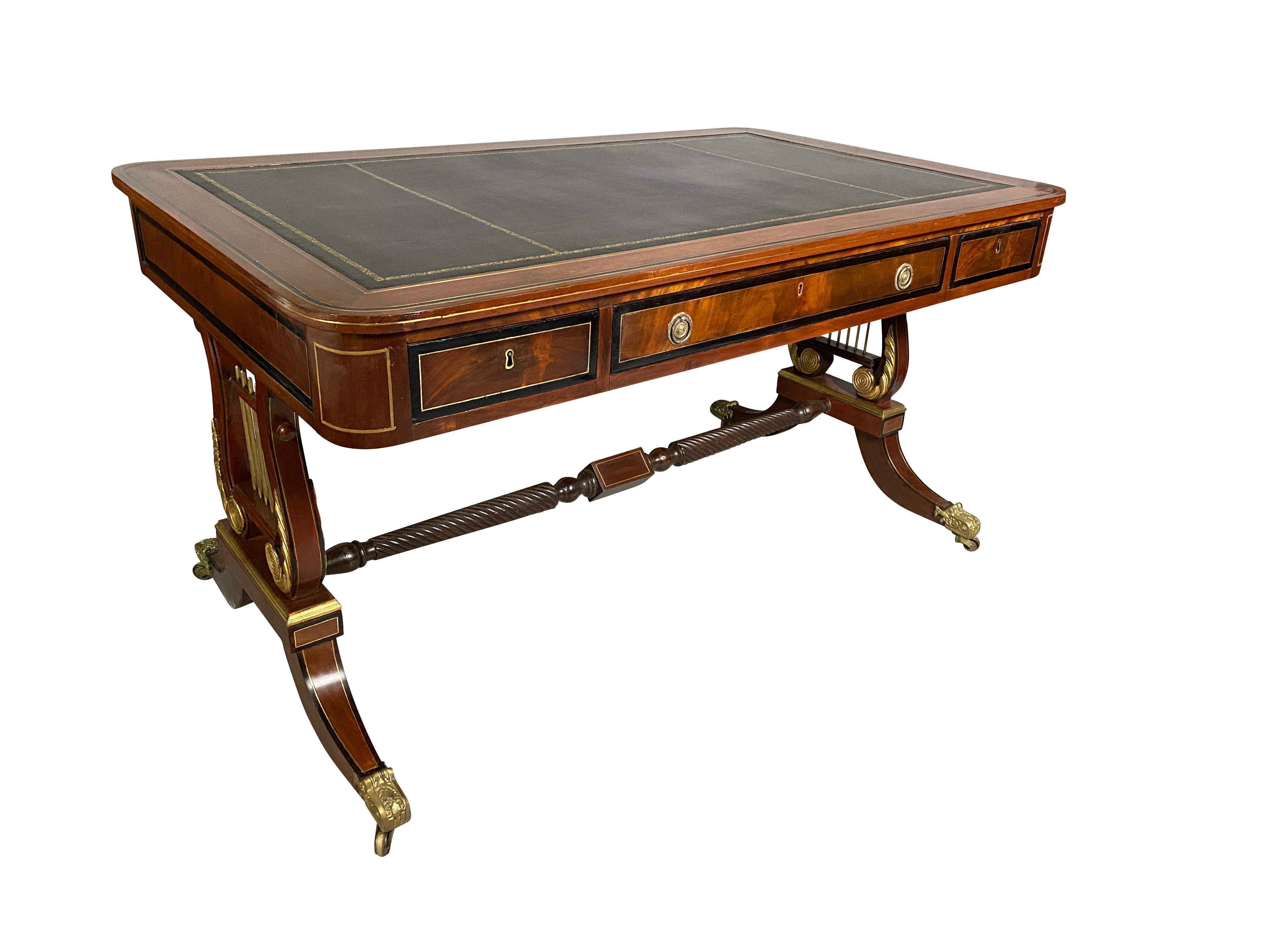 Rectangular black tooled leather top set within a border with brass and ebony inlay over a long drawer and flanked by a single drawer, the back of the desk is finished all raised on a trestle form base with partially gilded lyre form ends, joined by
